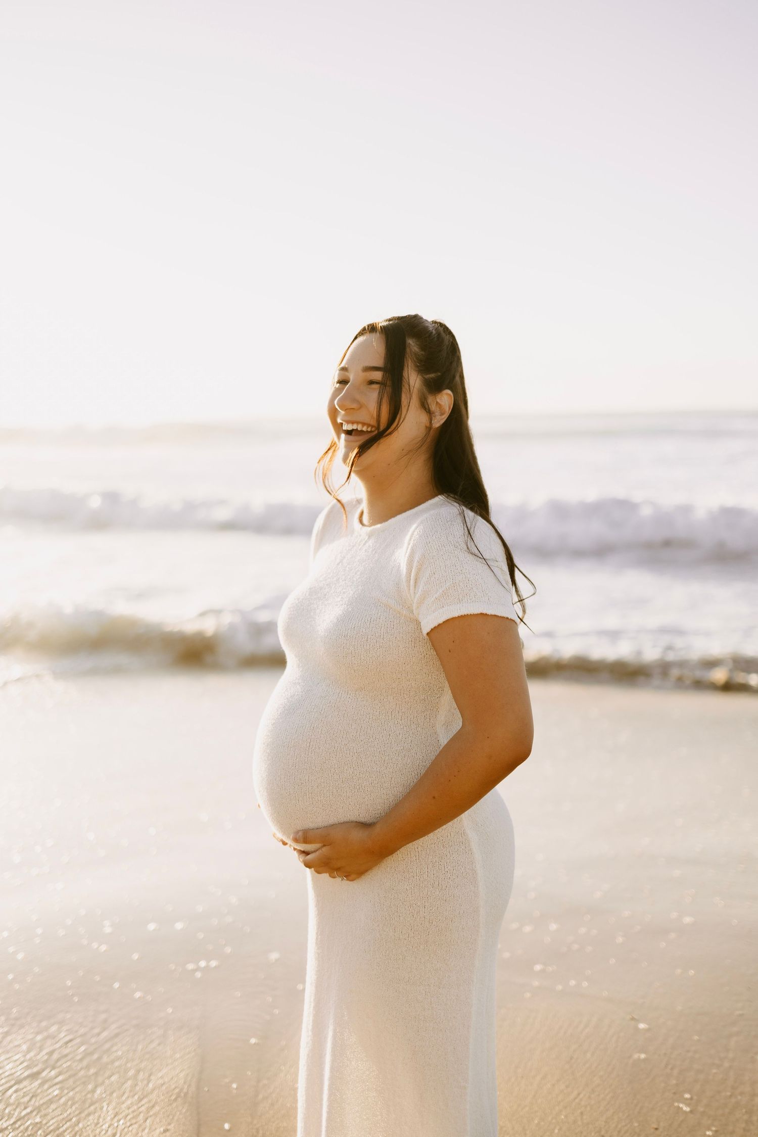 Smiling pregnant woman in white dress cradling baby bump on sunny beach with ocean waves behind her.