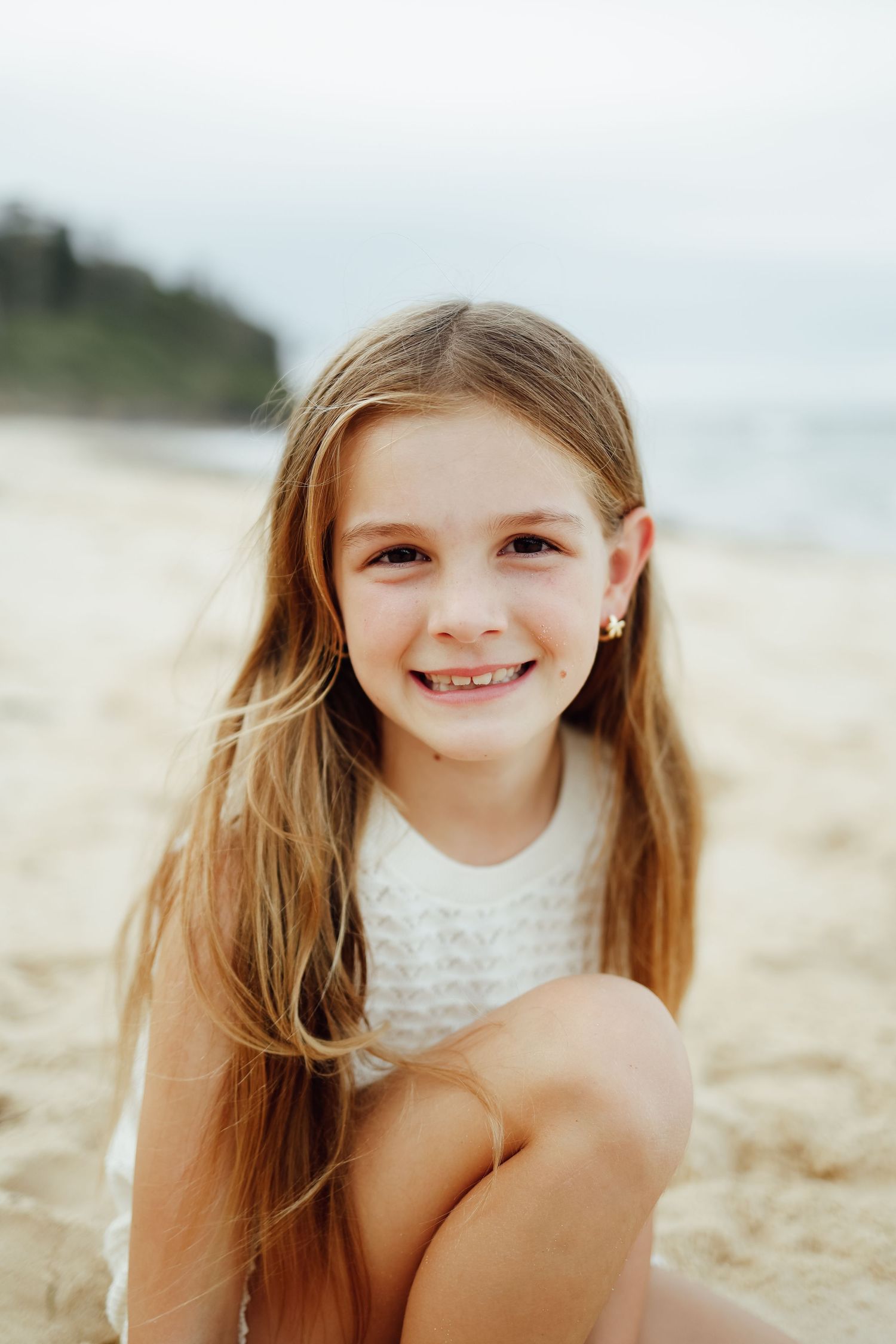 Smiling young girl with long blonde hair sitting on sandy beach wearing white dress.
