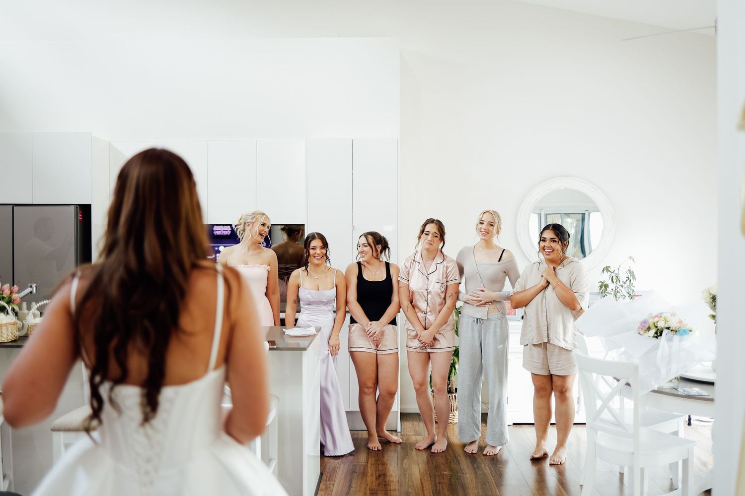 Bride reveals wedding dress to excited bridesmaids in bright white room during wedding morning preparations.