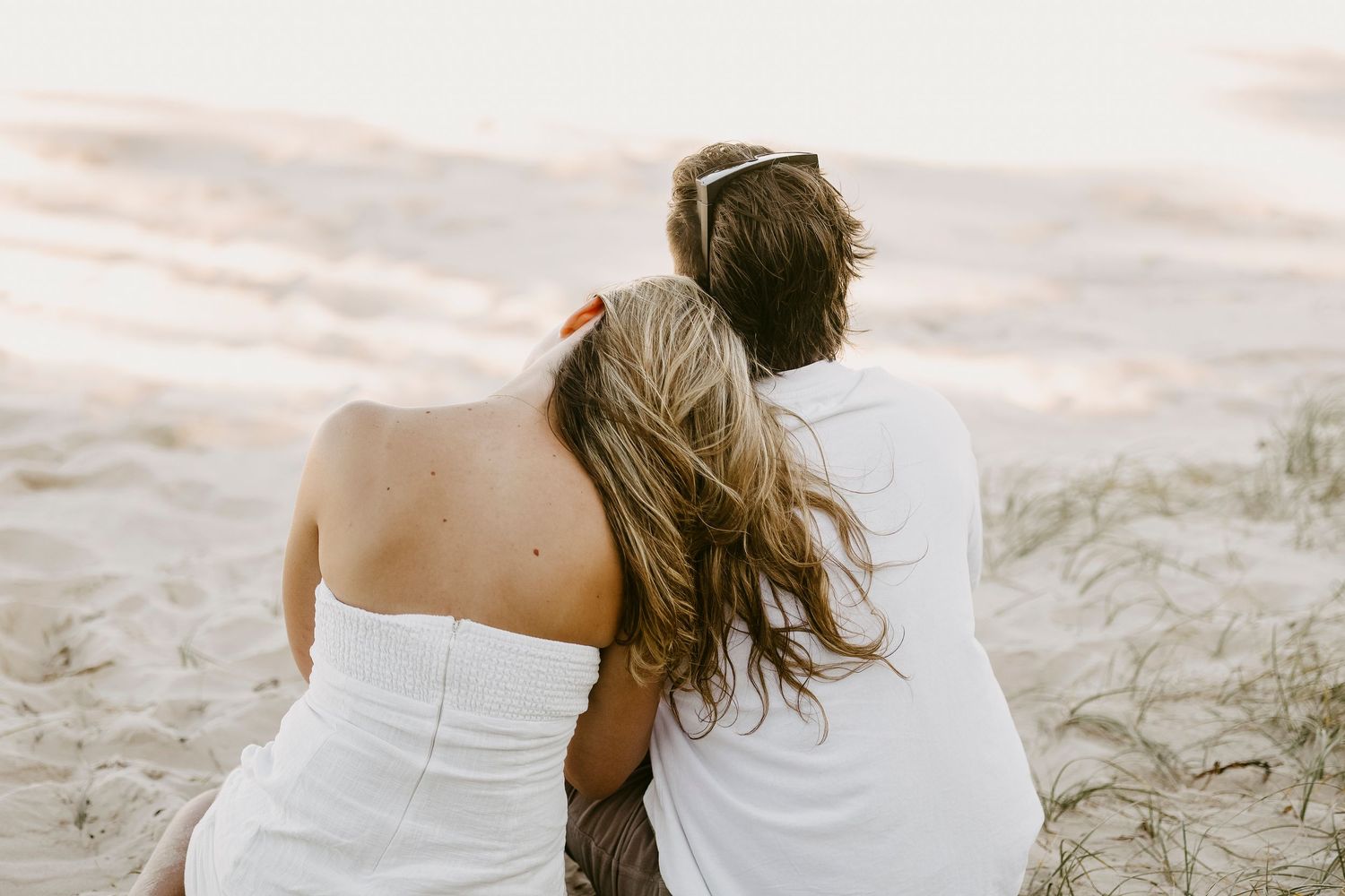 Romantic couple sitting together on sandy beach dunes at golden hour, woman leaning on man's shoulder.