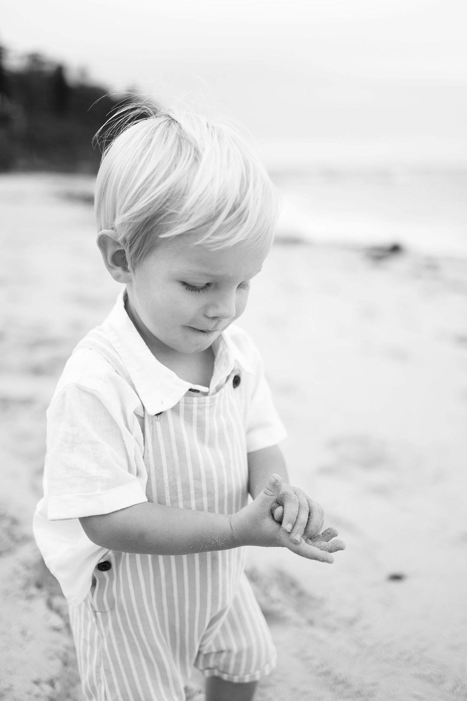 Black and white portrait of a toddler boy in striped overalls looking down on a beach.