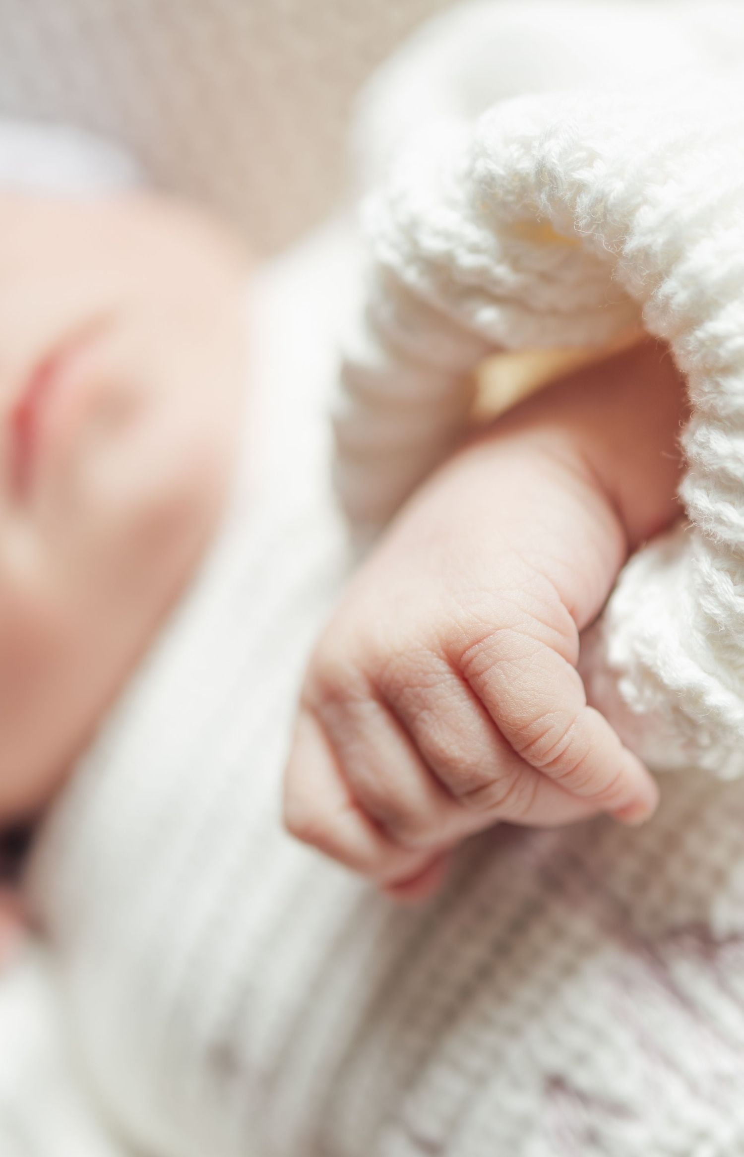 Close-up of a sleeping newborn baby's tiny hand and fingers, wrapped in a soft white knit blanket.