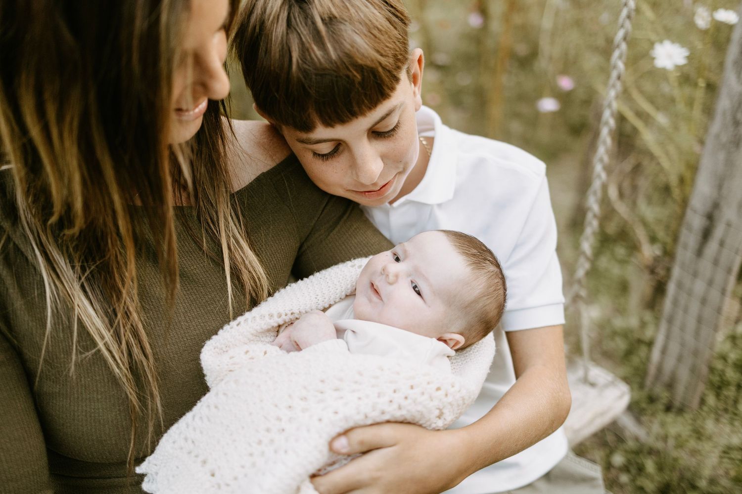 A mother and young boy lovingly gaze at a newborn baby wrapped in a white knit blanket outdoors.