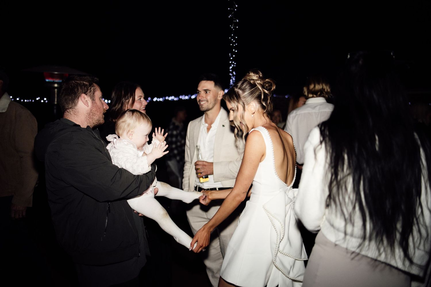 Couple in white attire dancing with guests at an outdoor evening engagement party.