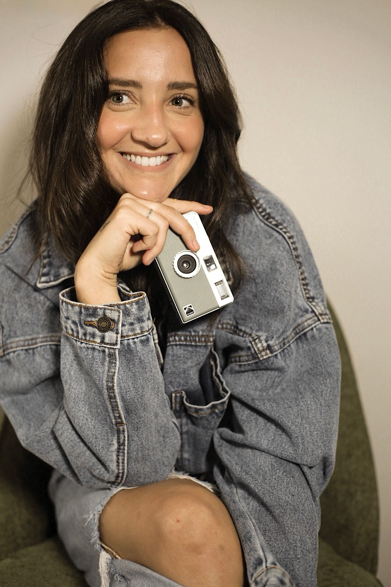 Smiling woman in denim jacket holding a vintage camera, posing casually with hand on chin. Nashville based photographer.
