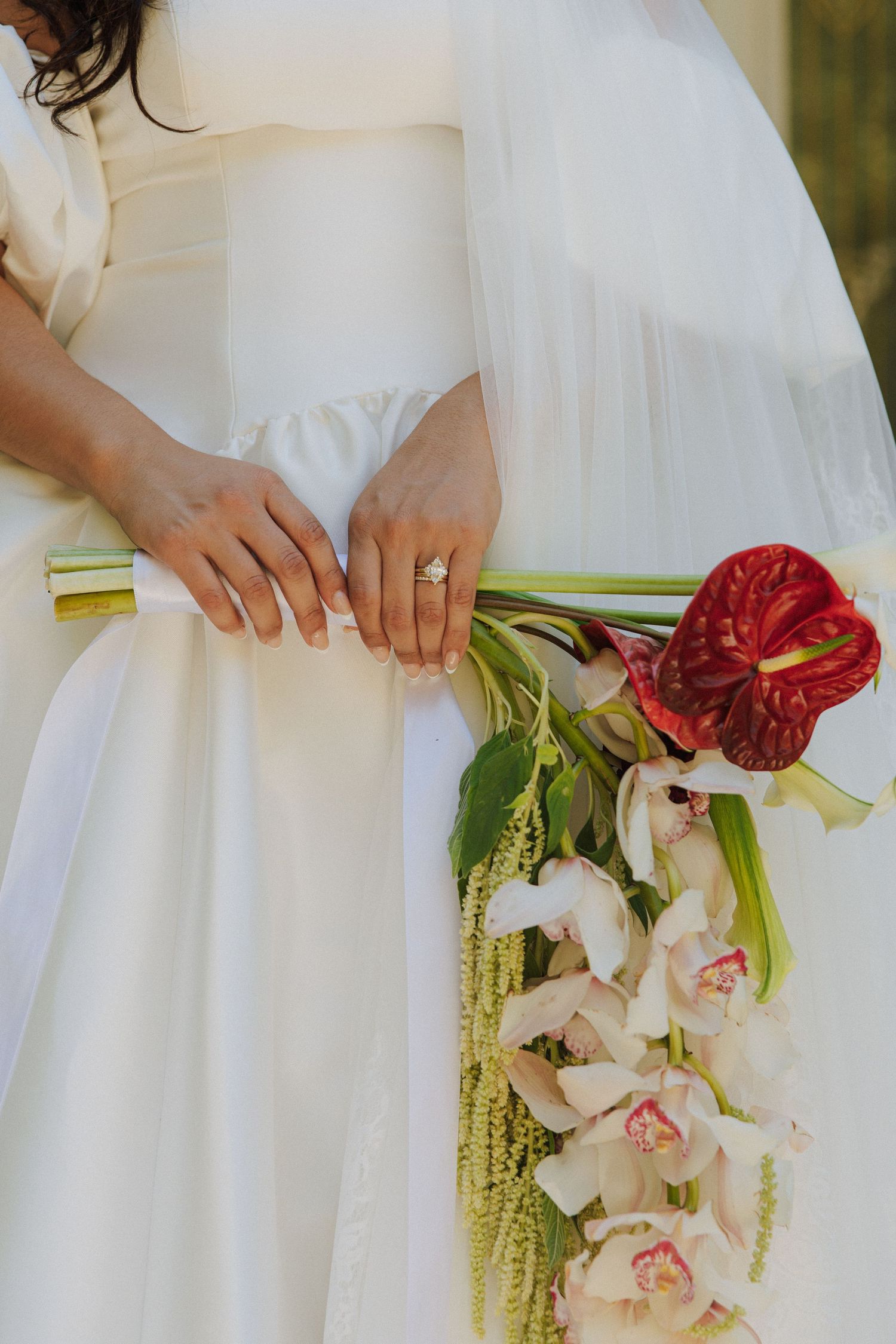 Bride in white wedding dress holding elegant bouquet with red anthurium and orchids, showcasing diamond engagement ring.