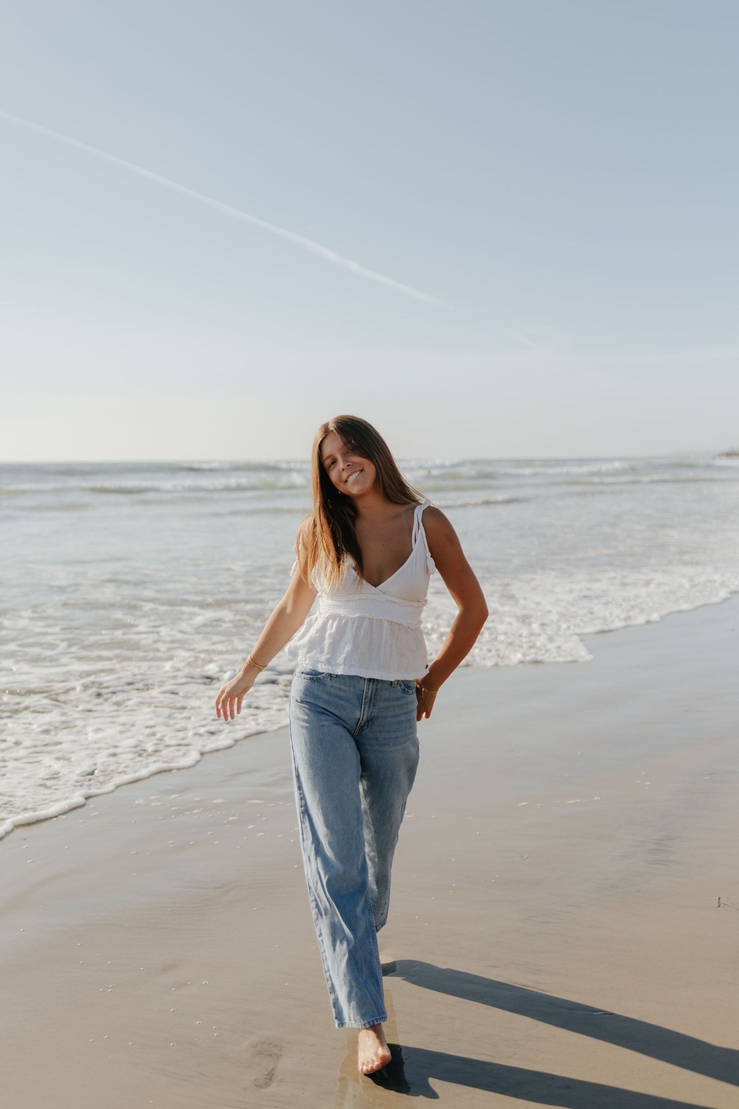A smiling young woman in a white top and jeans walks barefoot along a sunny ocean shoreline.