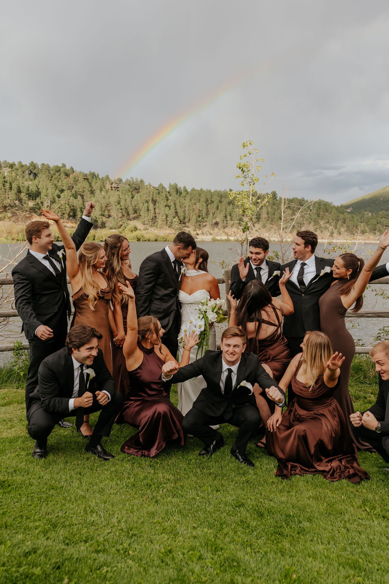 Wedding party celebrates with bride and groom kissing under a rainbow, bridesmaids in brown dresses and groomsmen in black suits.