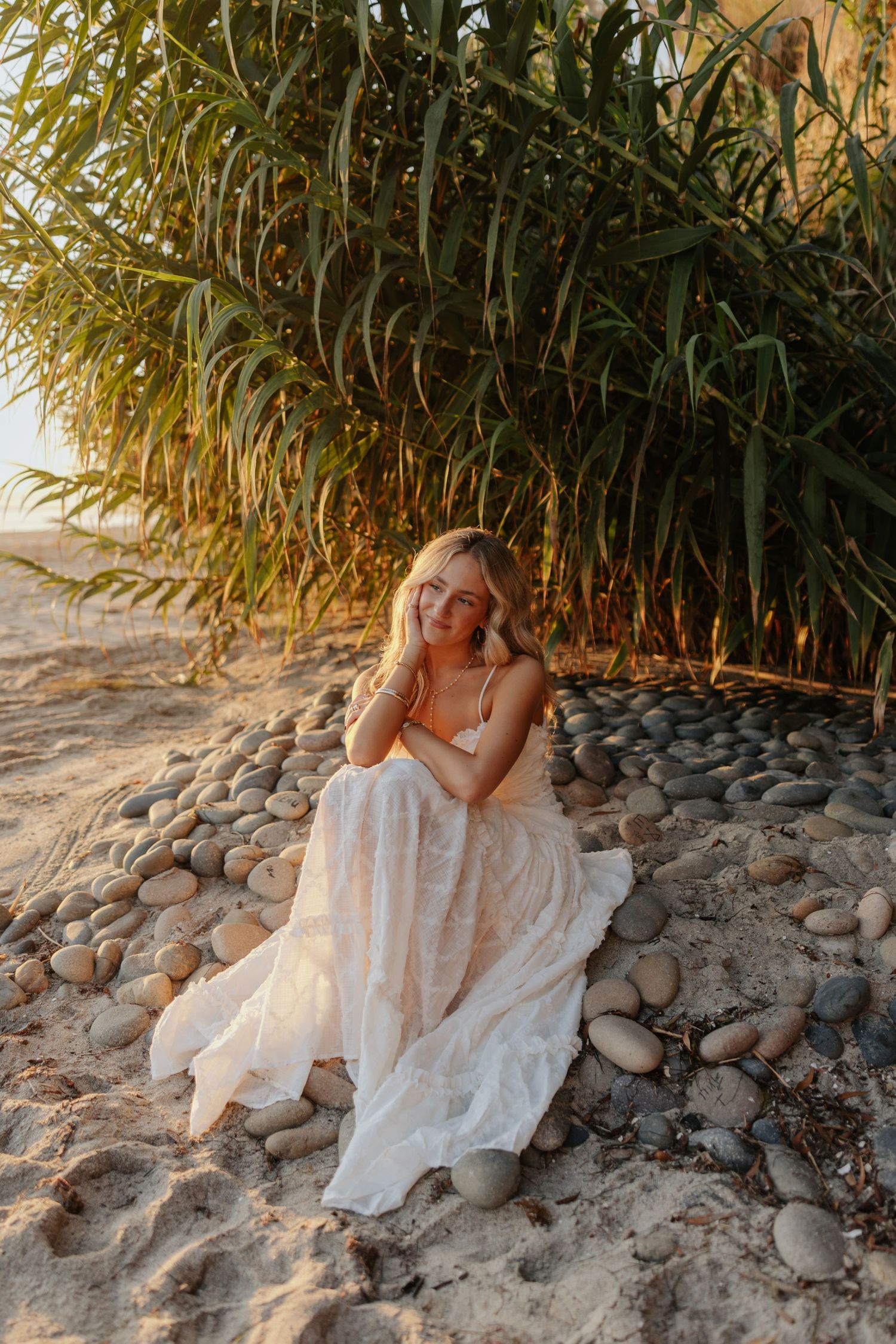 A woman in a white lace dress sits on pebbles by lush green reeds on a sandy beach at golden hour.