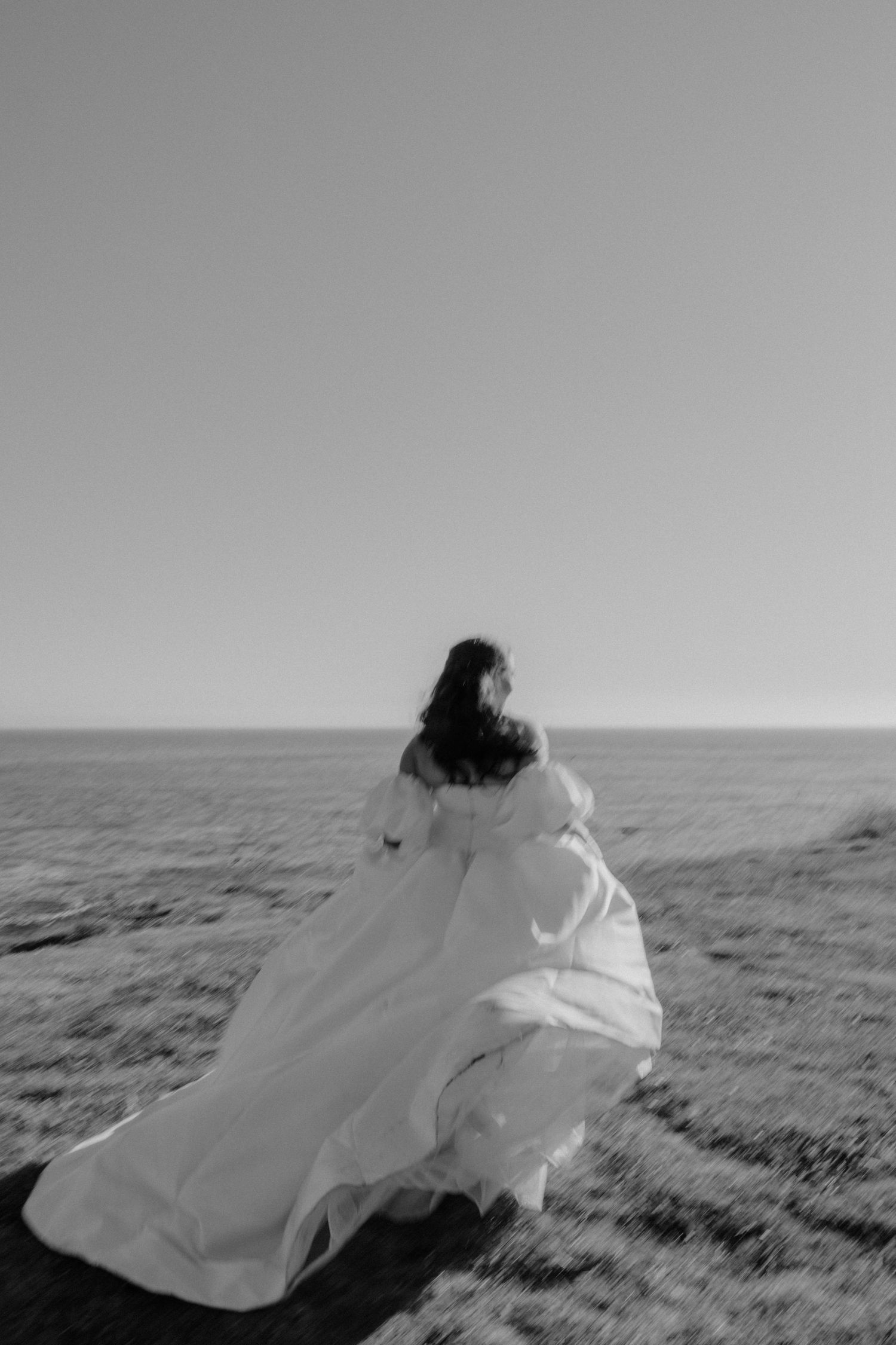 Black and white photo of a bride in a flowing white gown standing on a coastal cliff, her dress billowing in the wind.