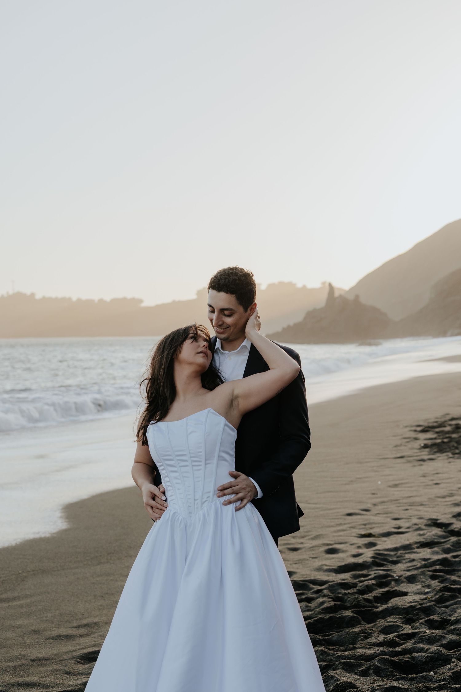 Bride and groom embrace romantically on a sandy beach with golden sunset and ocean waves.