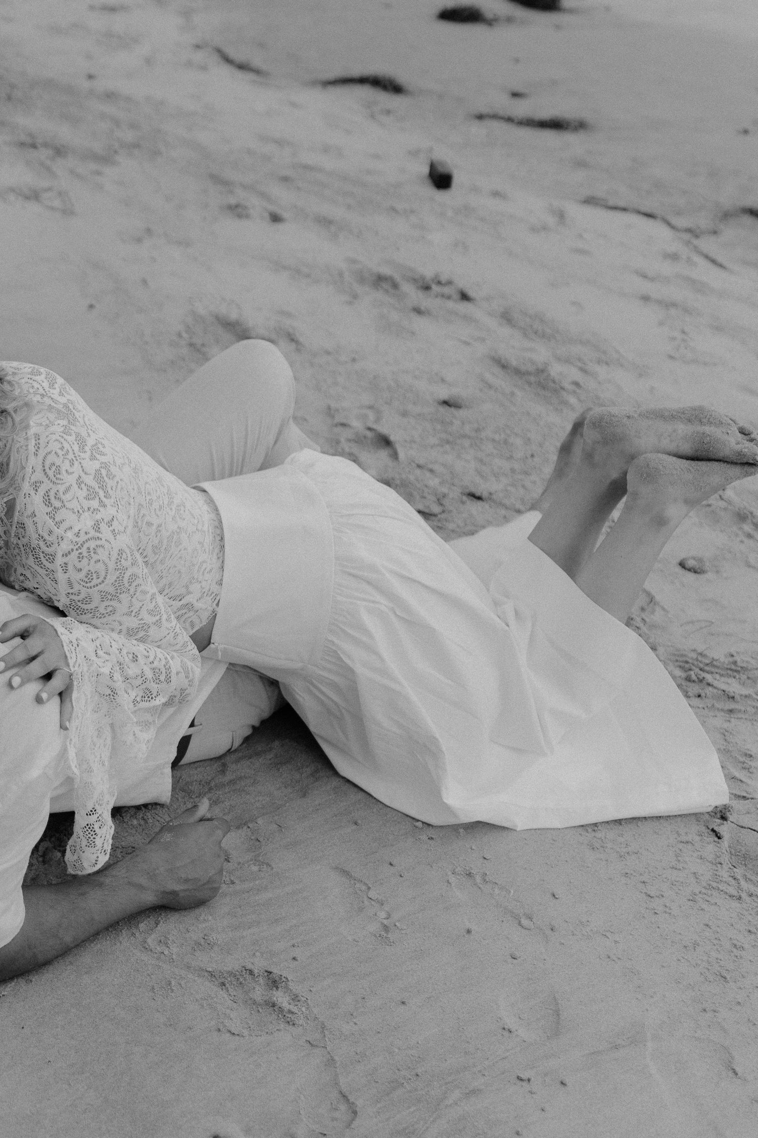 Black and white photo of couple lying together on sandy beach in white wedding attire.