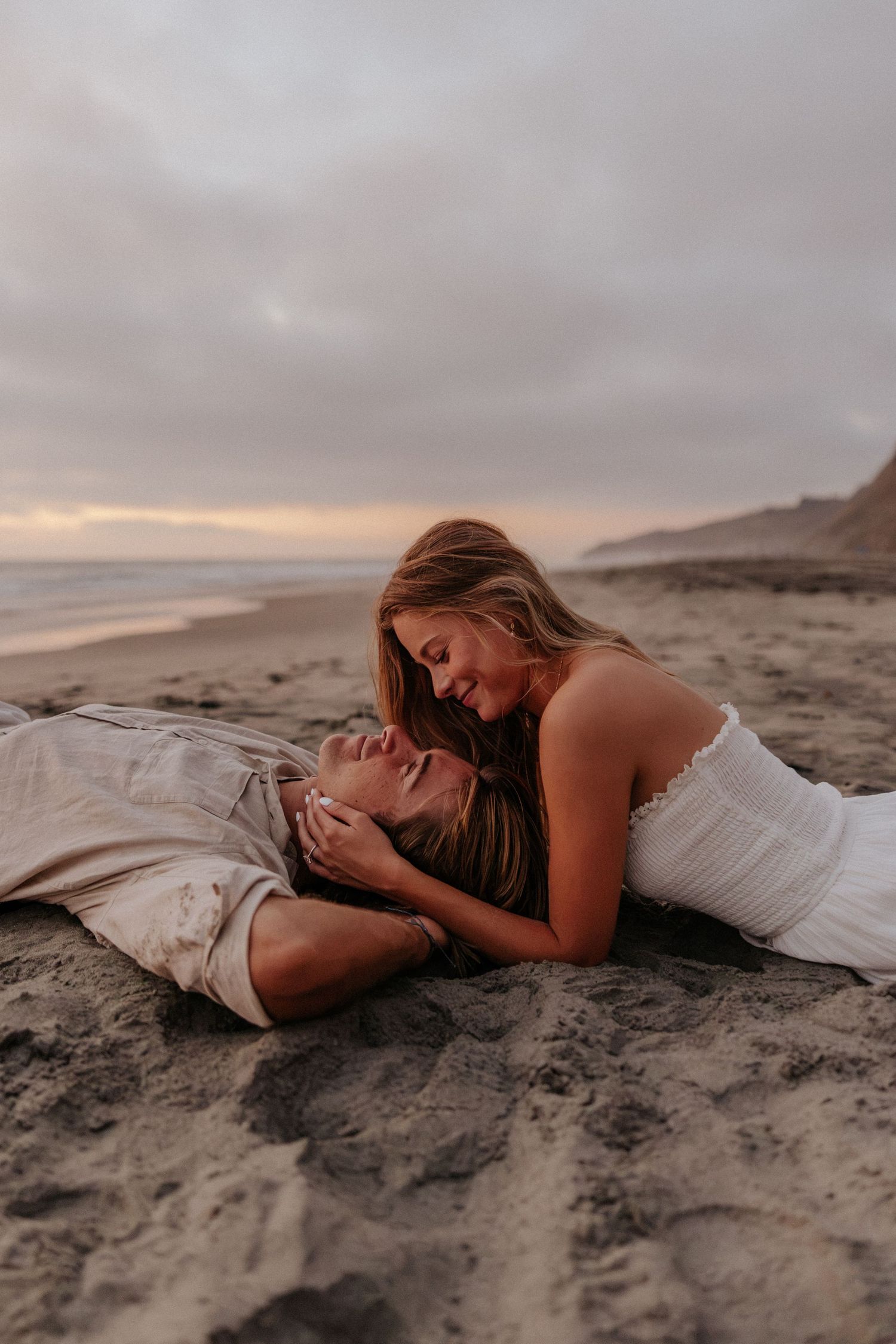 Couple sharing a romantic moment on a sandy beach at sunset, woman in white dress leaning over man lying in sand.