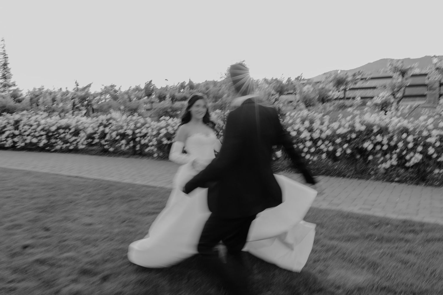 Black and white photo of bride and groom playfully running together on a manicured lawn.