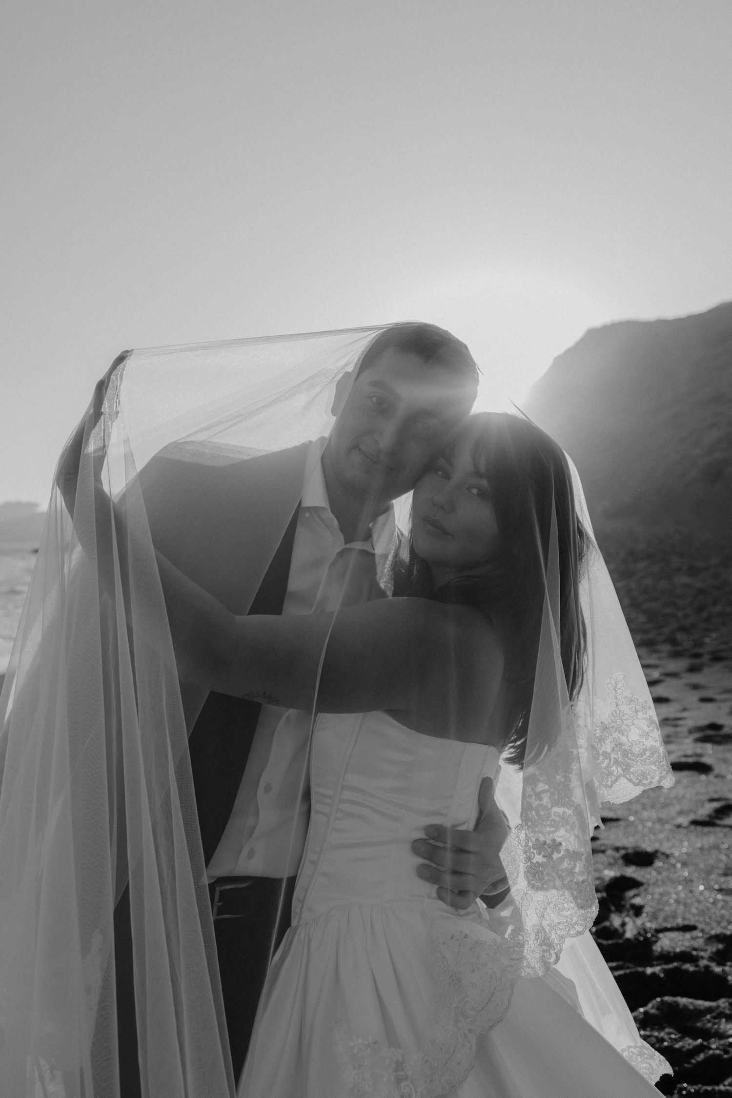 Black and white beach wedding photo of couple embracing under veil with sunset backdrop.