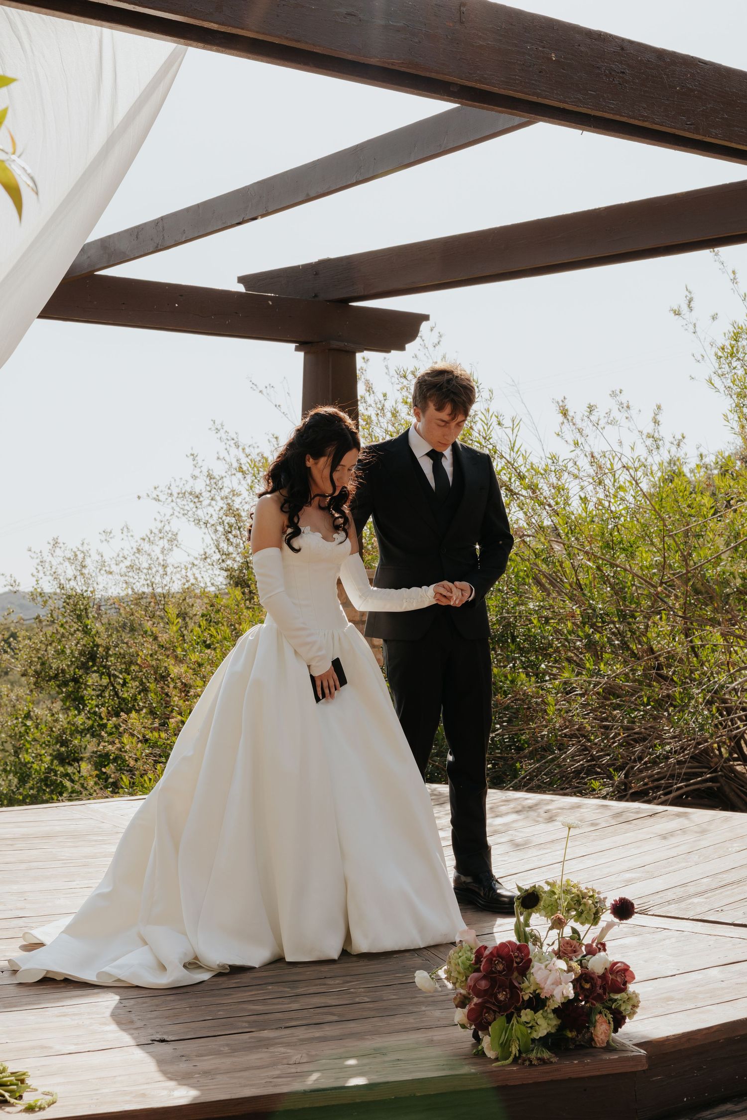 Bride in off-shoulder ball gown and groom in black suit holding hands under a wooden pergola.