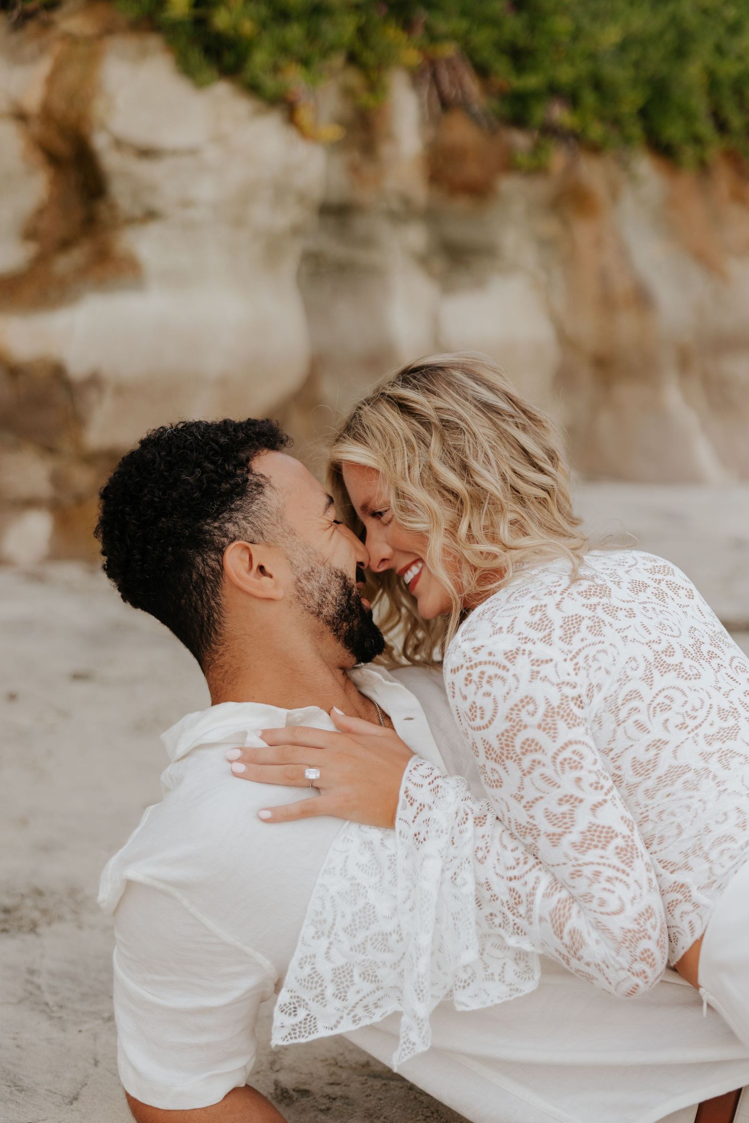 Smiling couple shares intimate almost-kiss, bride in white lace dress with engagement ring visible.