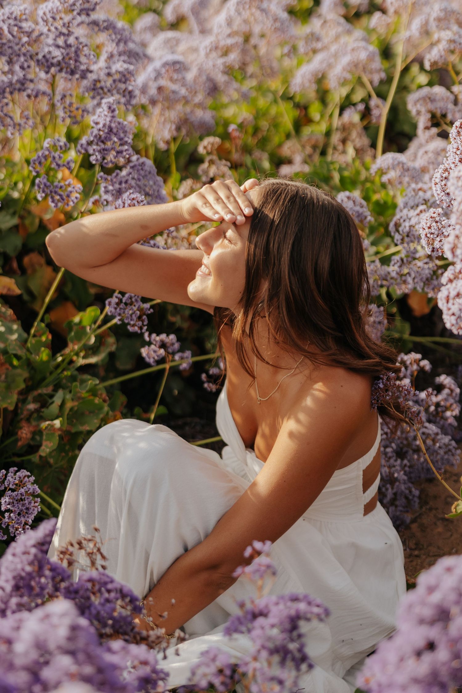 Brunette woman in white dress sitting among purple wildflowers shielding eyes from bright sunlight.