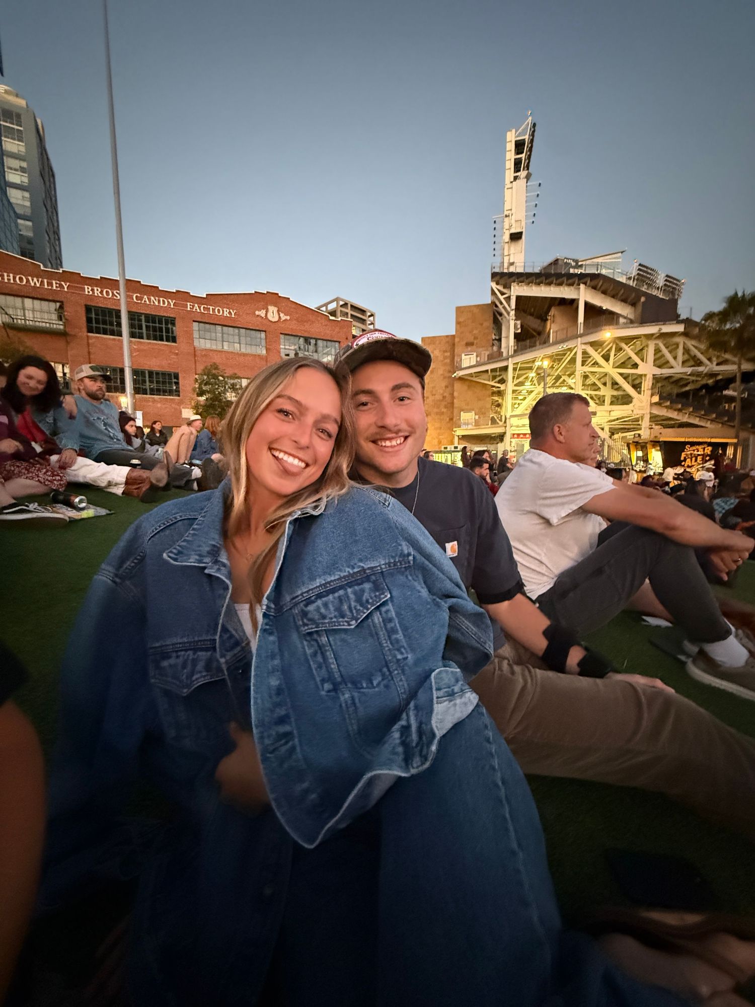 Smiling couple in denim jackets sitting on grass at an outdoor venue during golden hour.