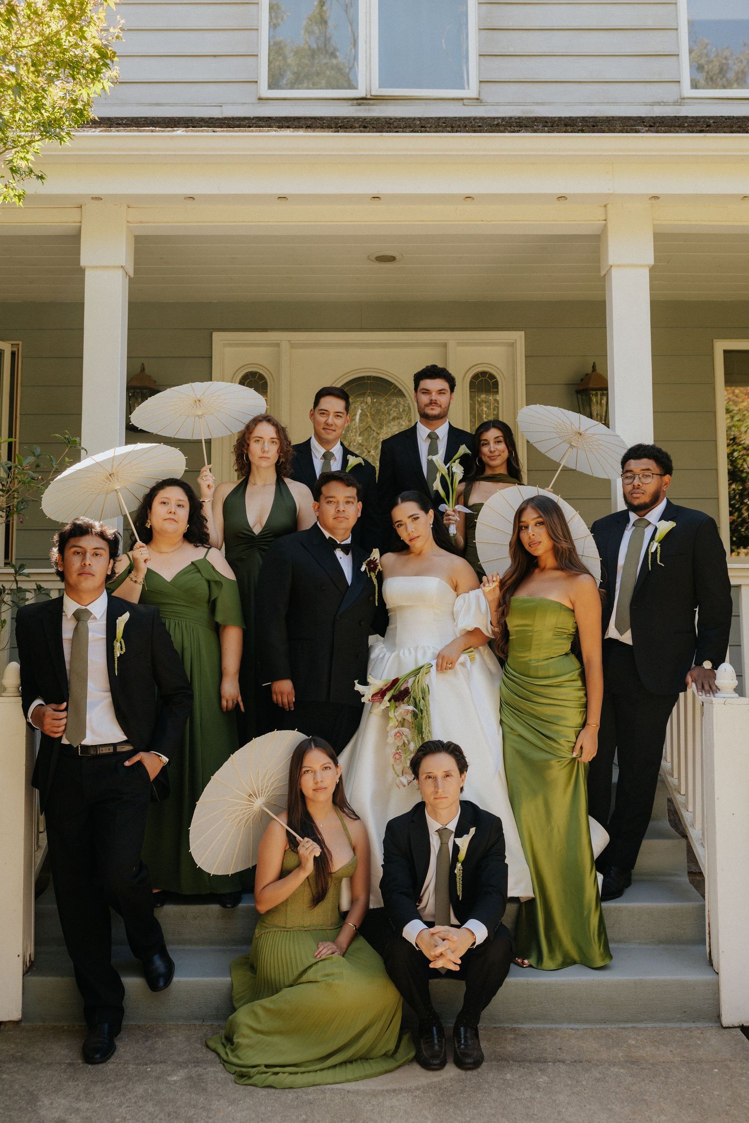 Wedding party poses on porch steps in green satin gowns, tuxedos, and white parasols with bride and groom centered.