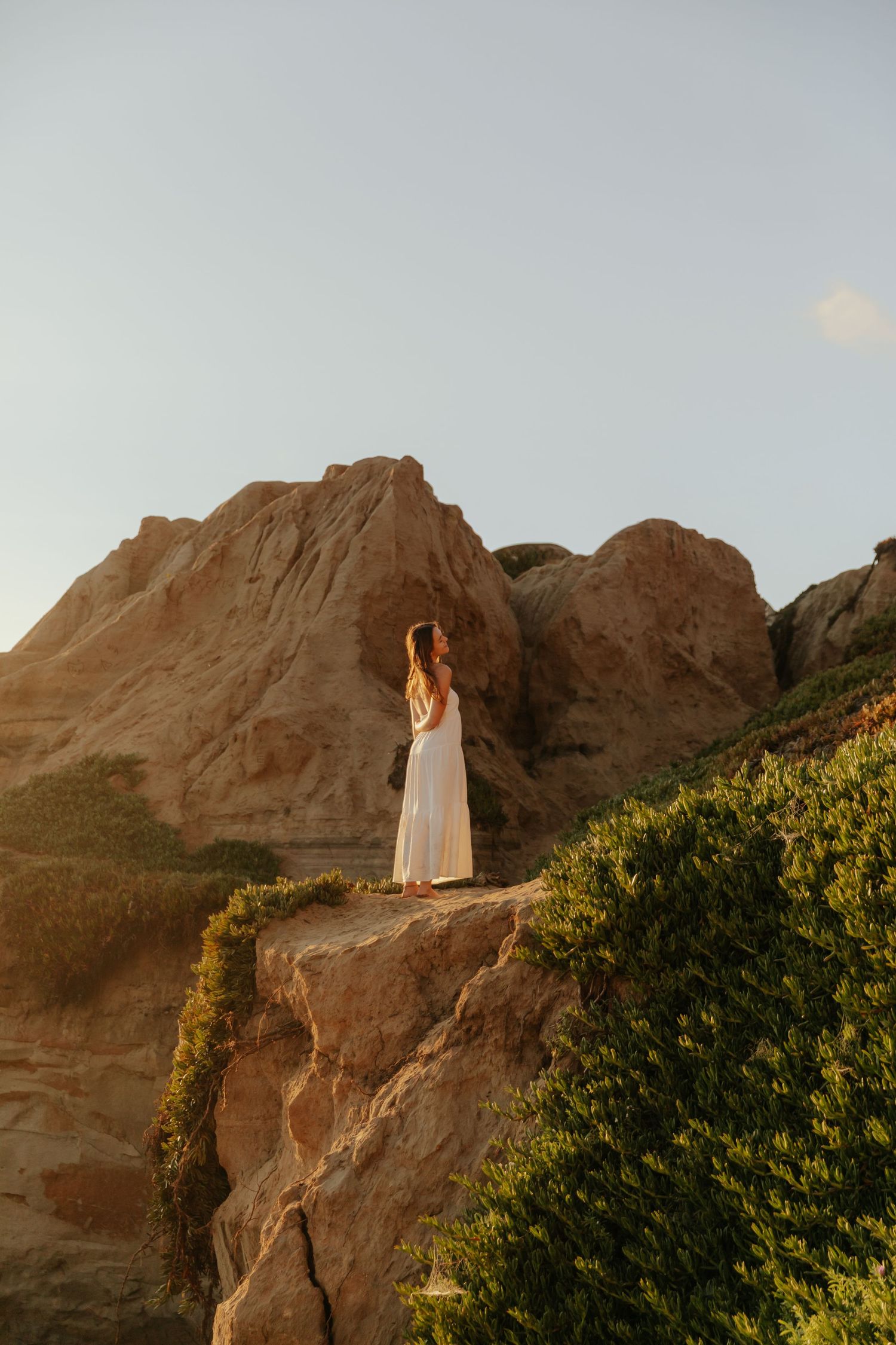 Woman in white dress stands on rocky coastal cliffs at golden hour with mountains behind her.