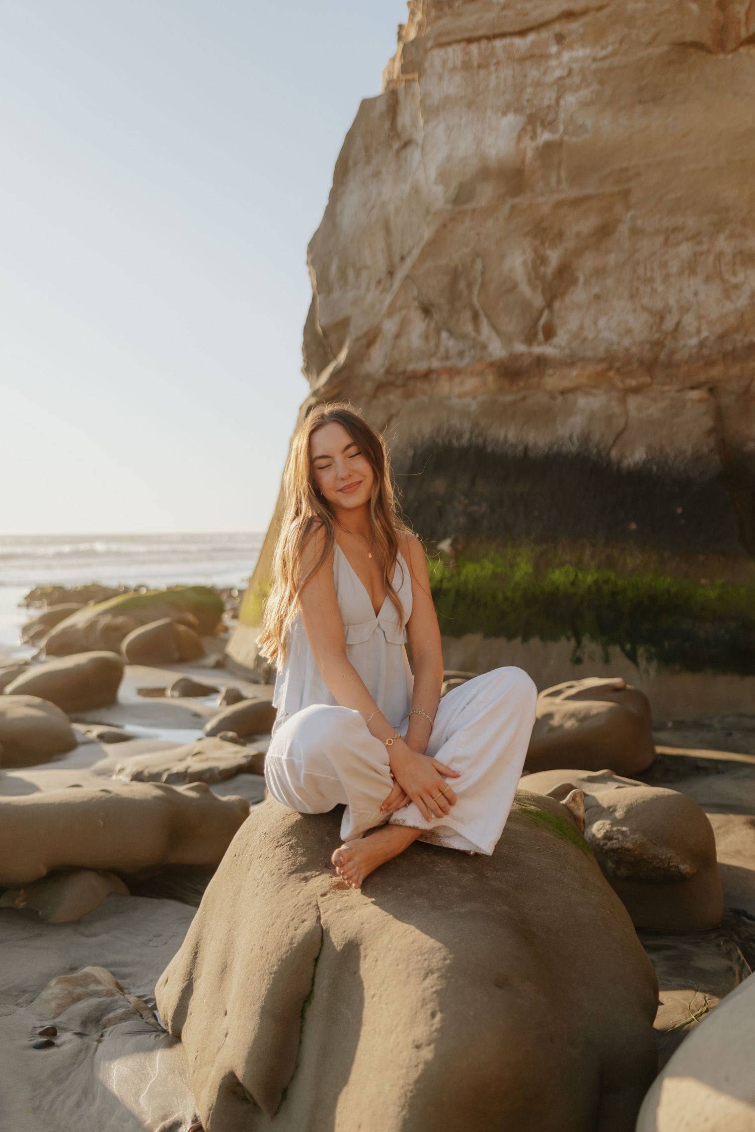 Smiling woman in white outfit sitting cross-legged on beach rocks near sandstone cliffs at golden hour.