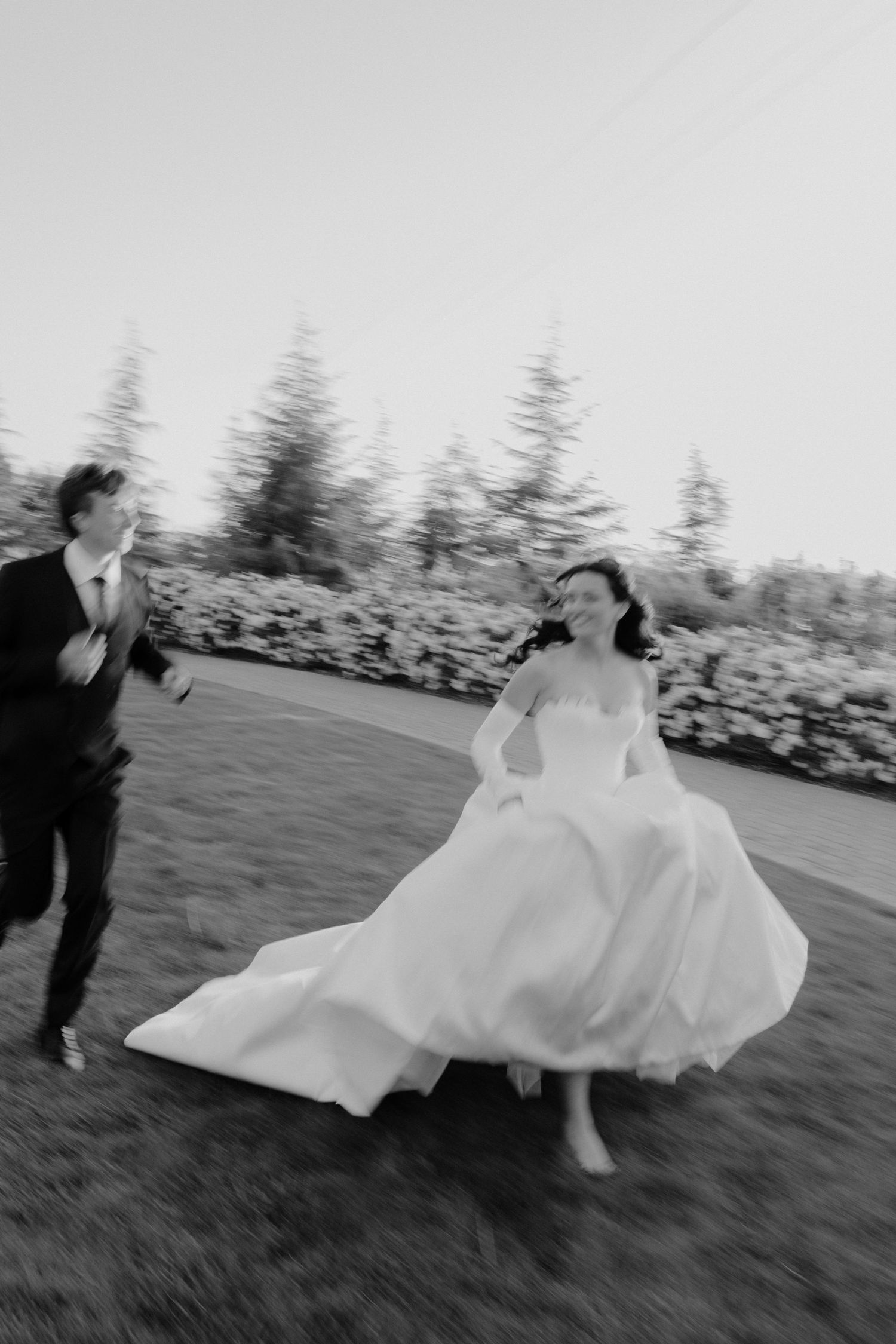Black and white motion blur photo of laughing bride and groom running together across garden lawn.
