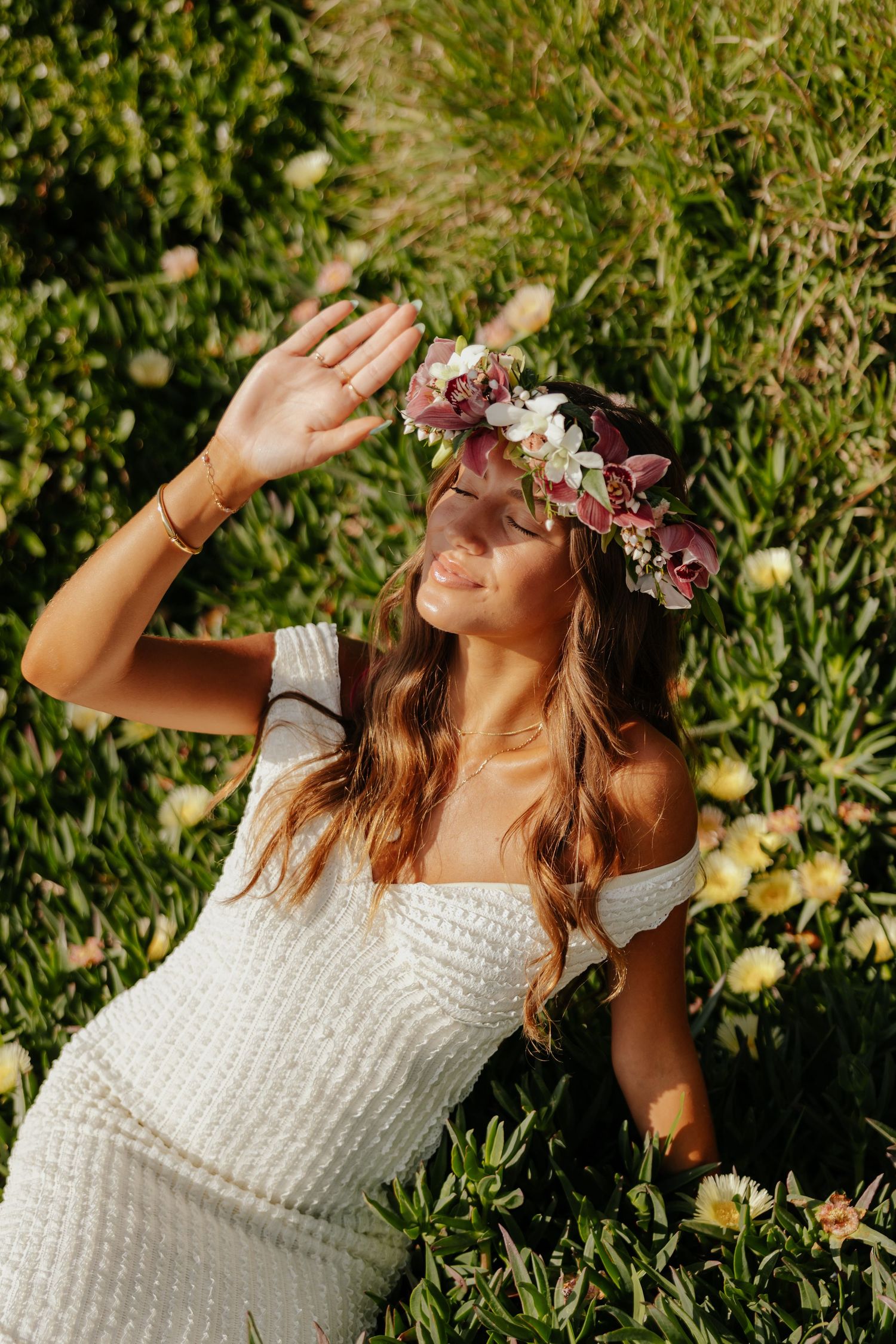 Bride wearing floral crown sitting in green meadow shielding eyes from golden sunlight.