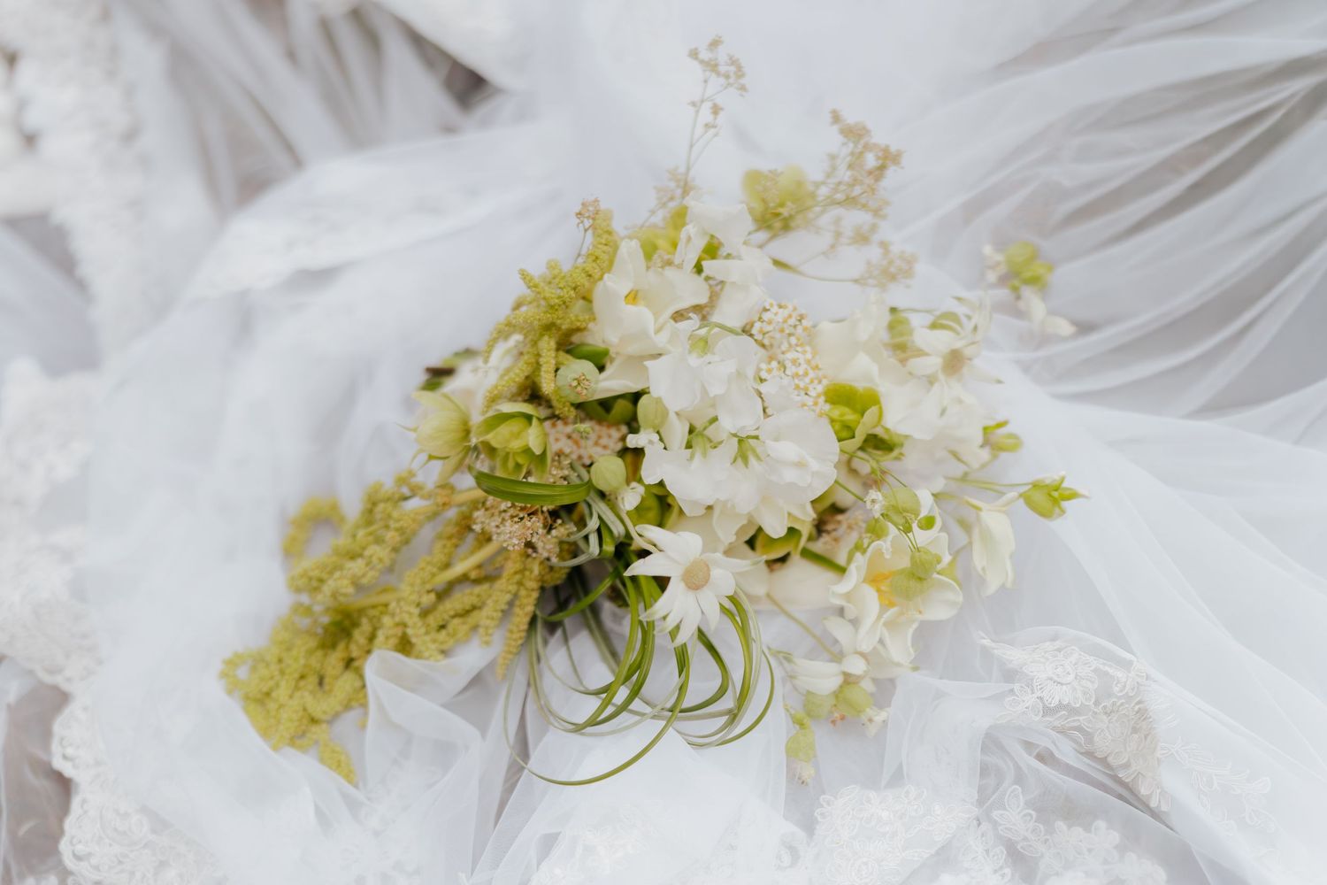 Elegant white and green wedding bouquet with wildflowers resting on a lace bridal veil.