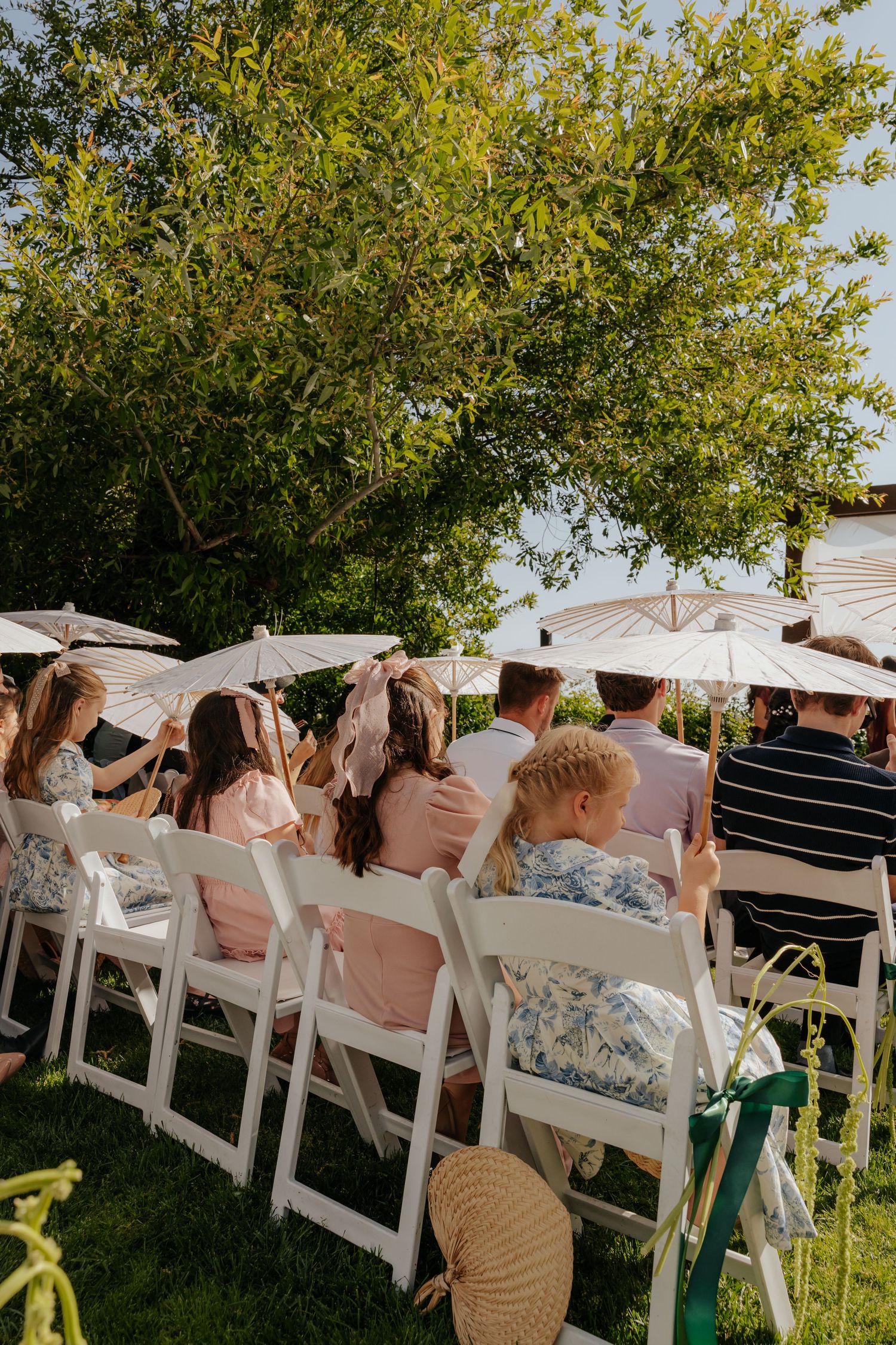 Wedding guests seated in white chairs under parasols at an outdoor garden ceremony on sunny day.
