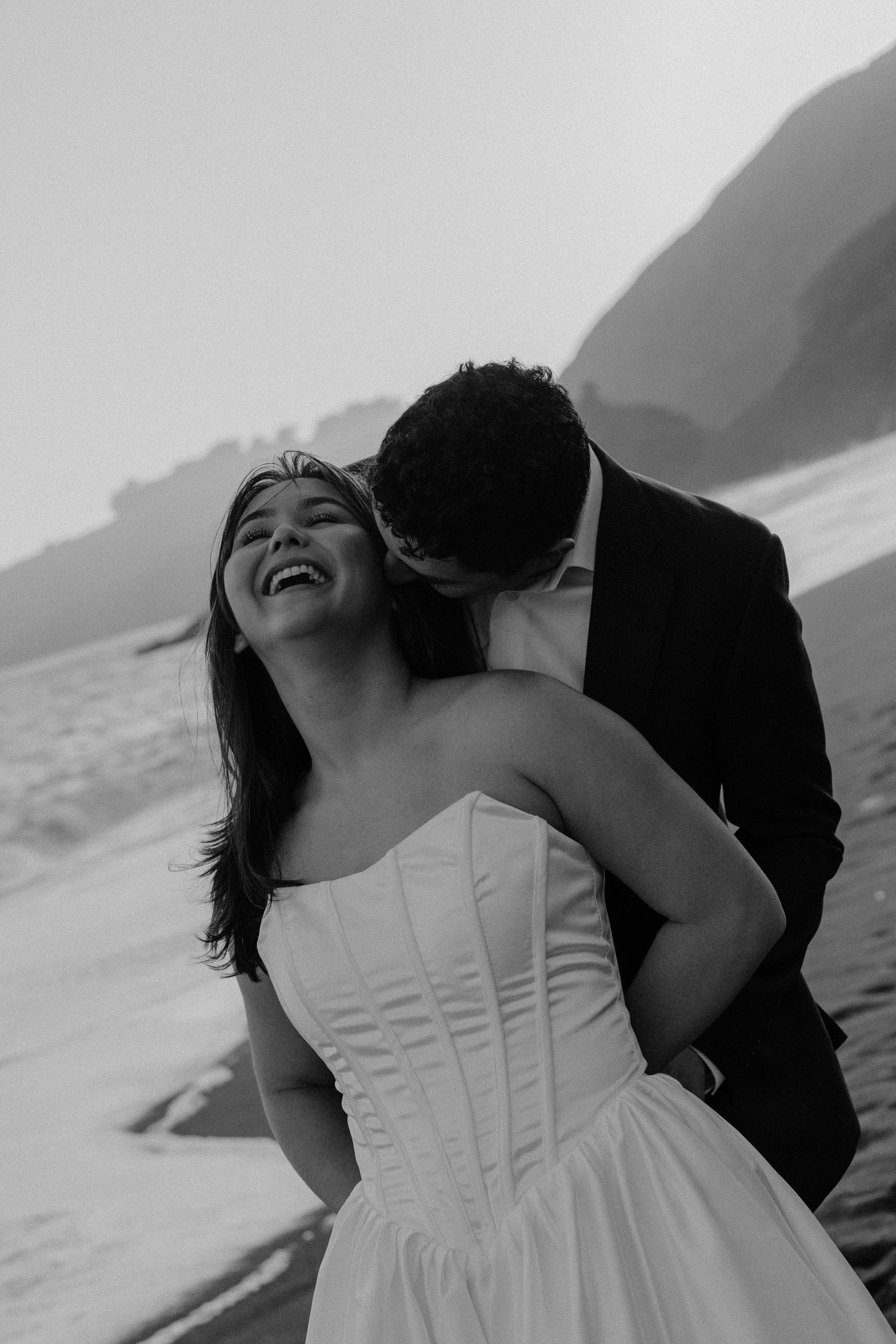 Black and white photo of laughing bride and groom sharing a tender beach moment at dusk.