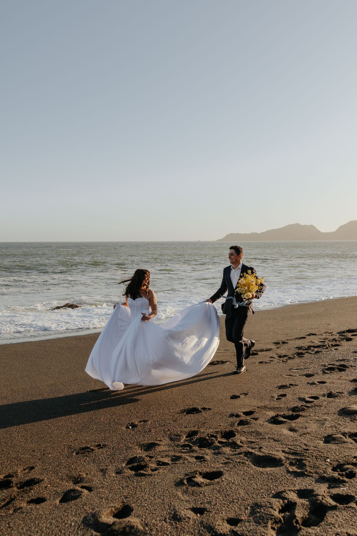 Bride and groom walk along sandy beach at sunset, groom holding a yellow flower bouquet.