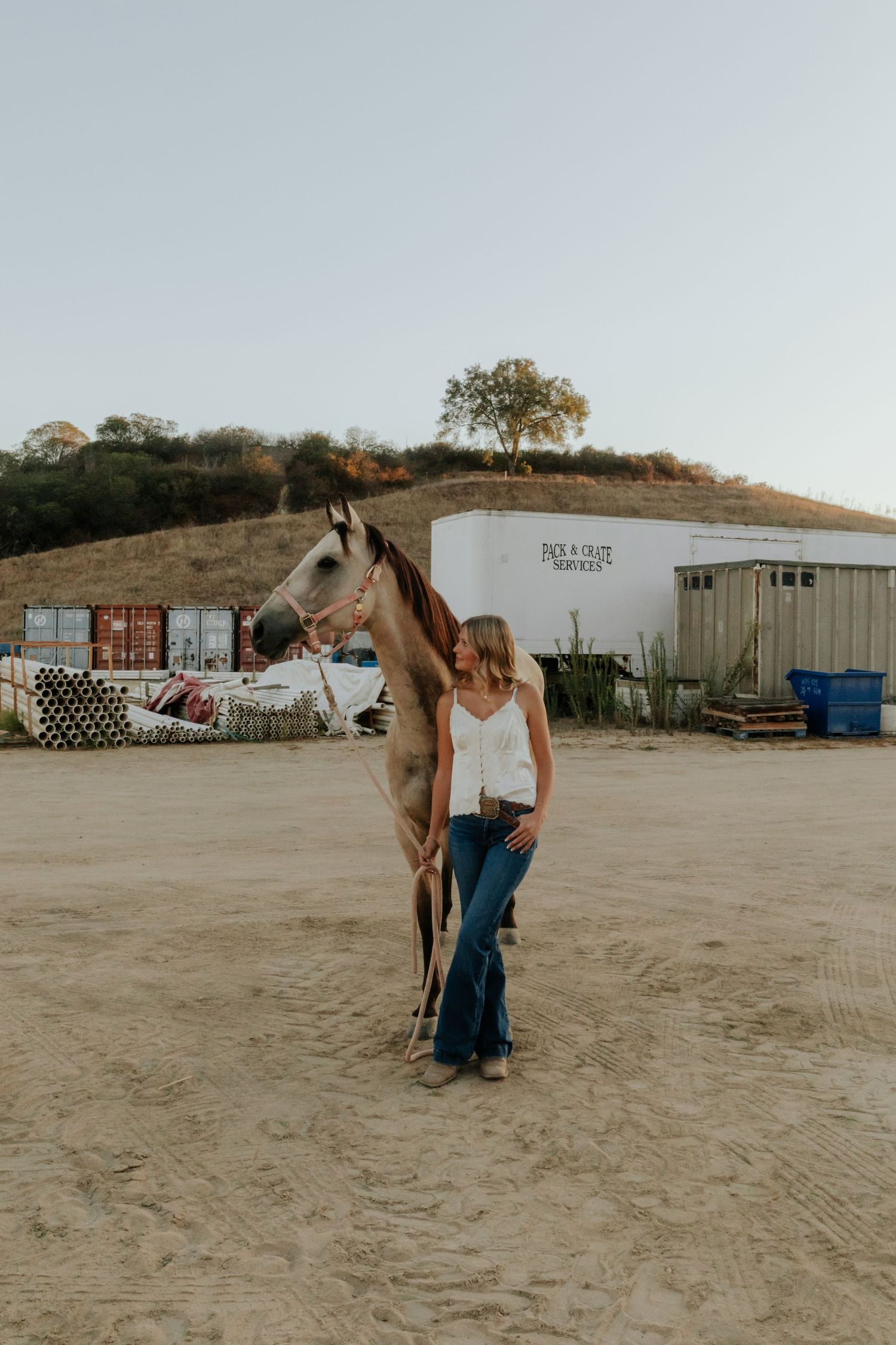 Woman in white top and jeans stands beside a horse on a sandy ranch with hills in the background.