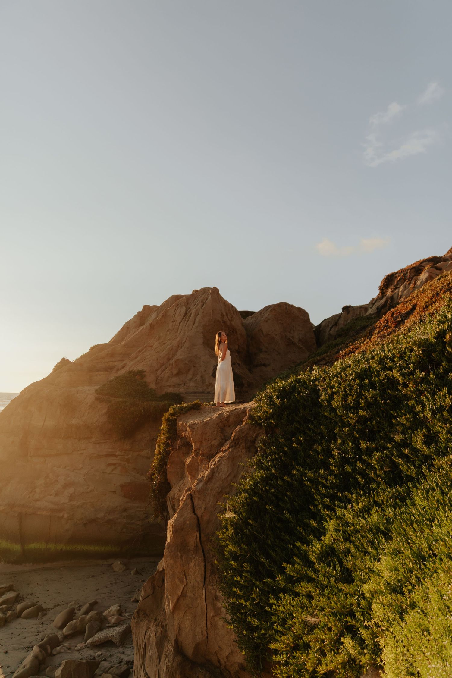 Bride in flowing white gown posing on rocky coastal cliffs bathed in golden sunset light.