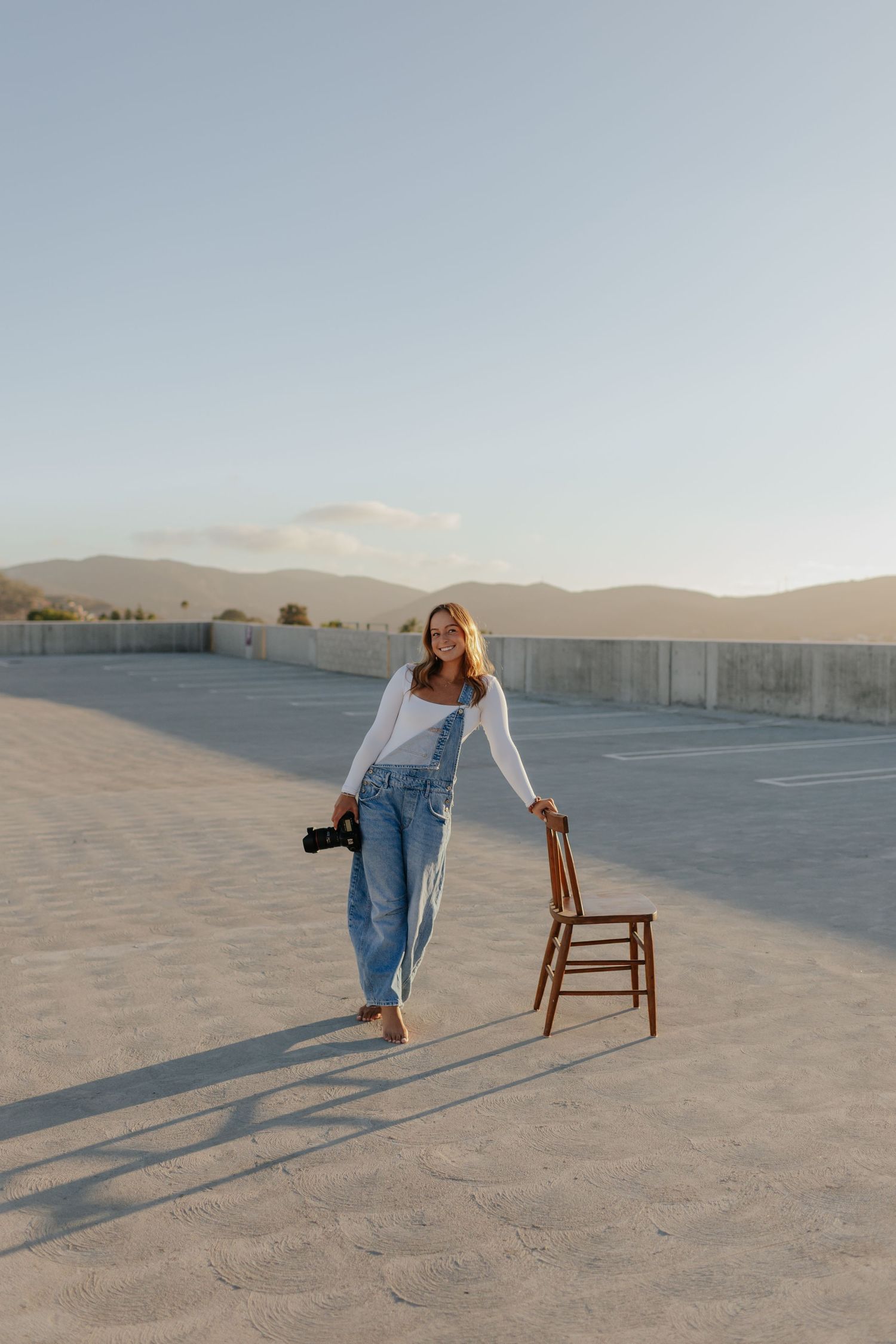 A smiling woman in denim overalls holds a camera and wooden chair on a sandy parking lot with mountains at sunset.