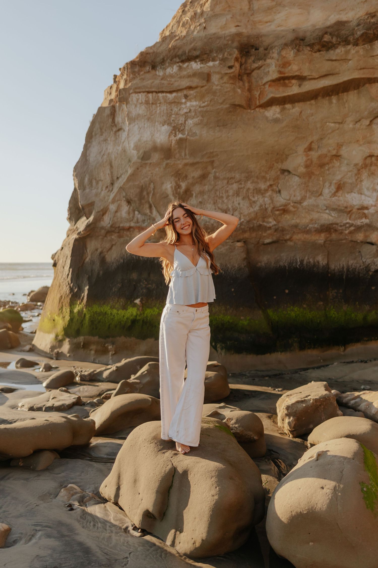Woman in white linen outfit standing on coastal rocks with dramatic sandstone cliffs at sunset.