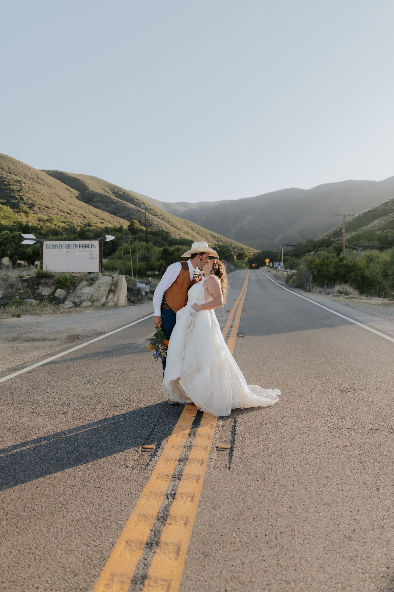 A bride and groom kissing on a rural road with scenic green mountains in the background.
