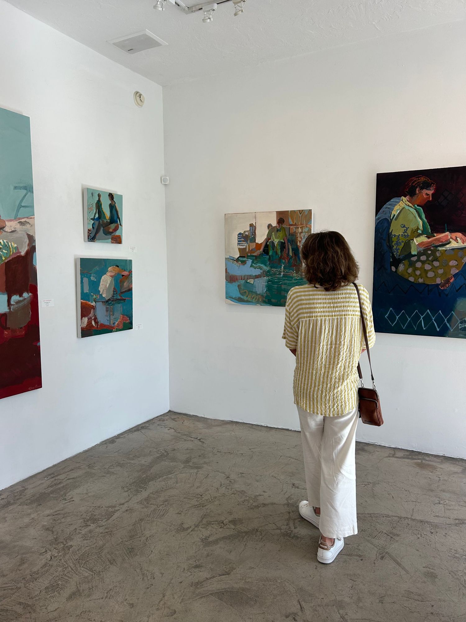 Woman in striped shirt viewing contemporary figurative paintings on white gallery walls.