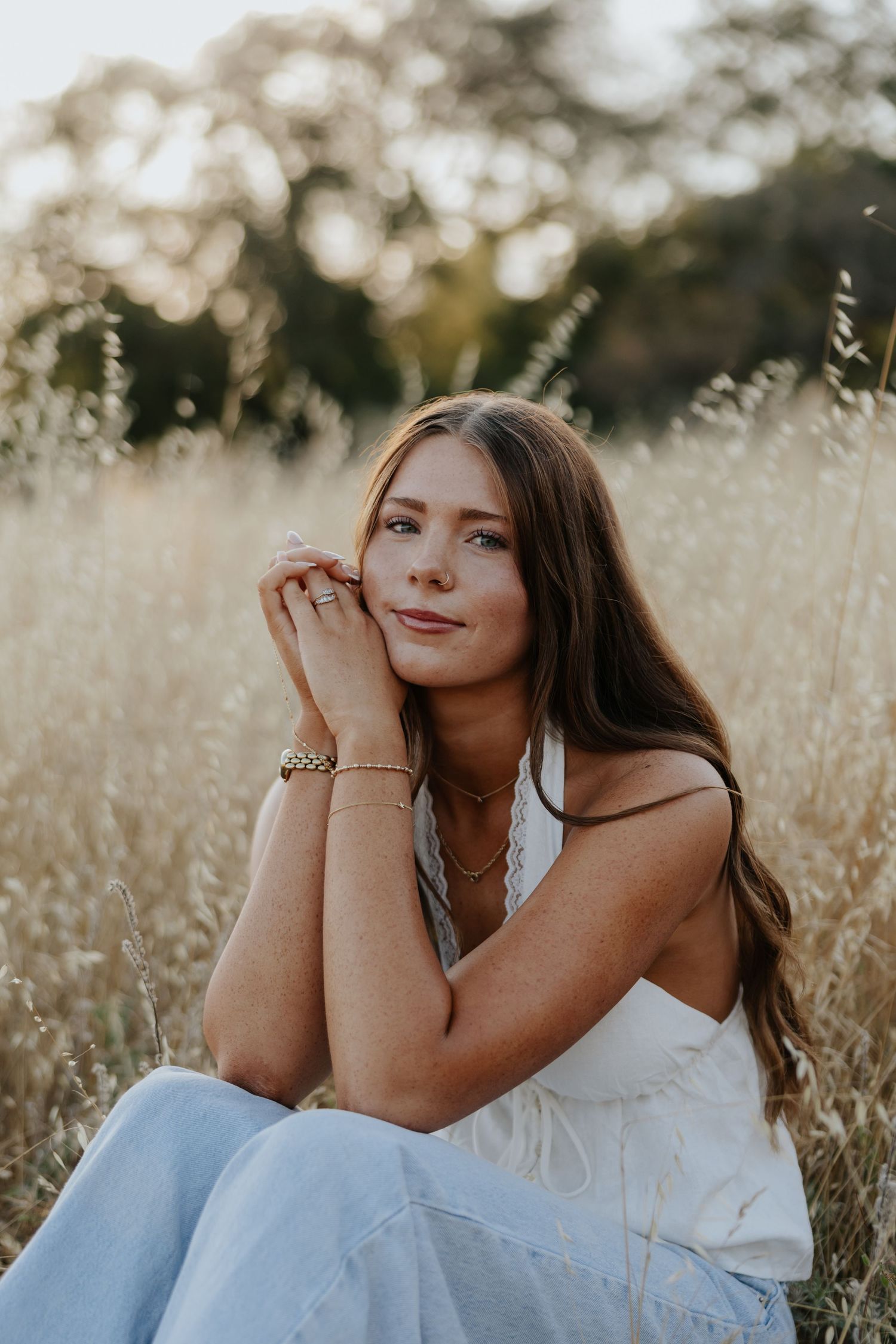 Young brunette woman in white dress sitting in golden field at sunset, hands clasped near face.