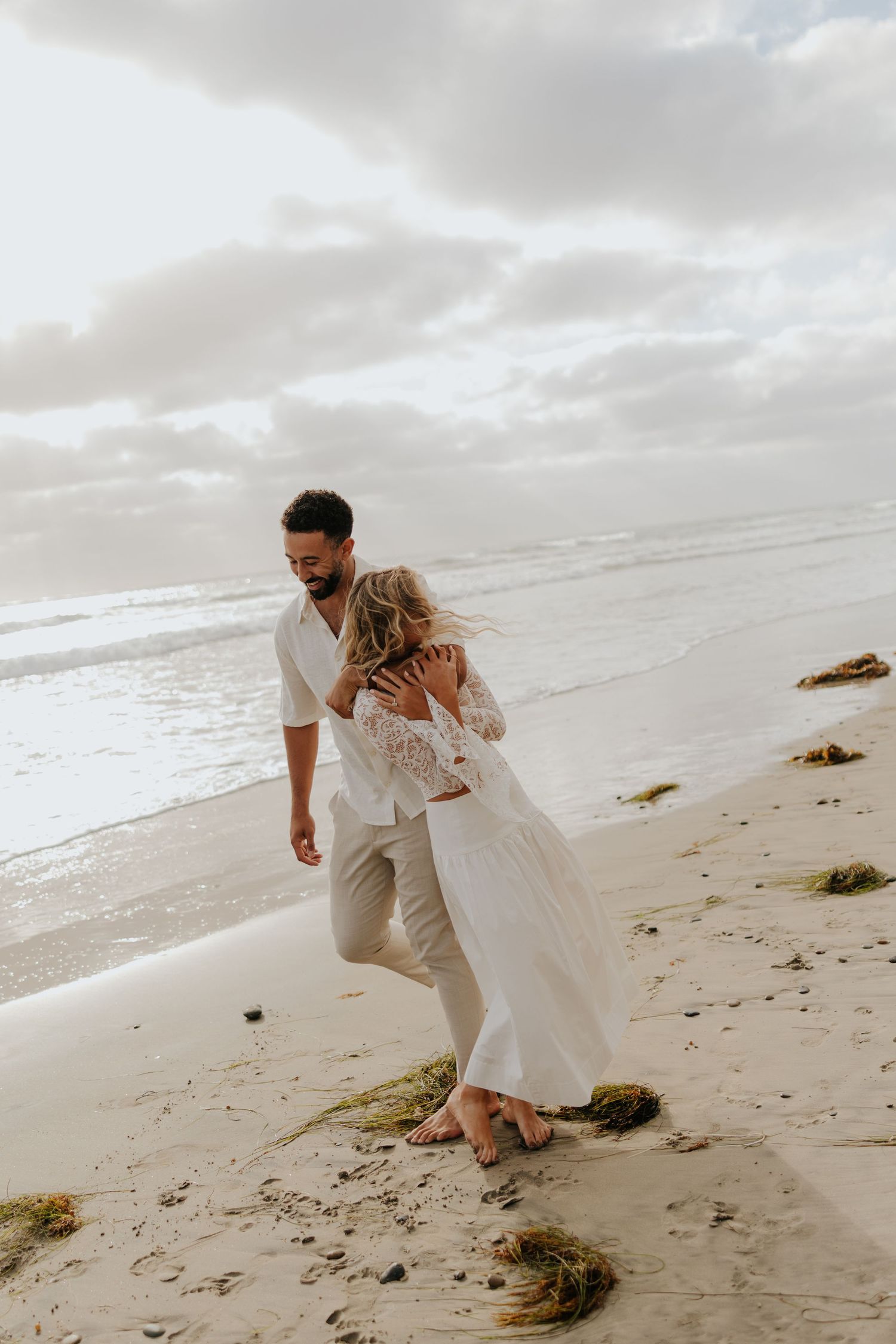 Couple in white wedding attire walking barefoot together on a sandy beach at sunset.