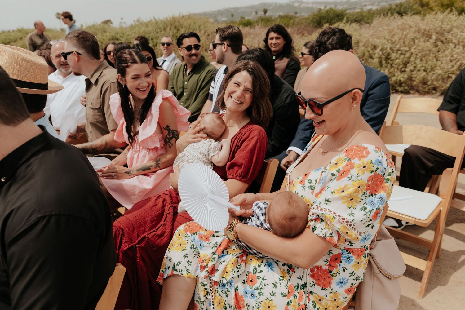 Wedding guests seated outdoors at coastal ceremony, including woman in floral dress holding infant baby.