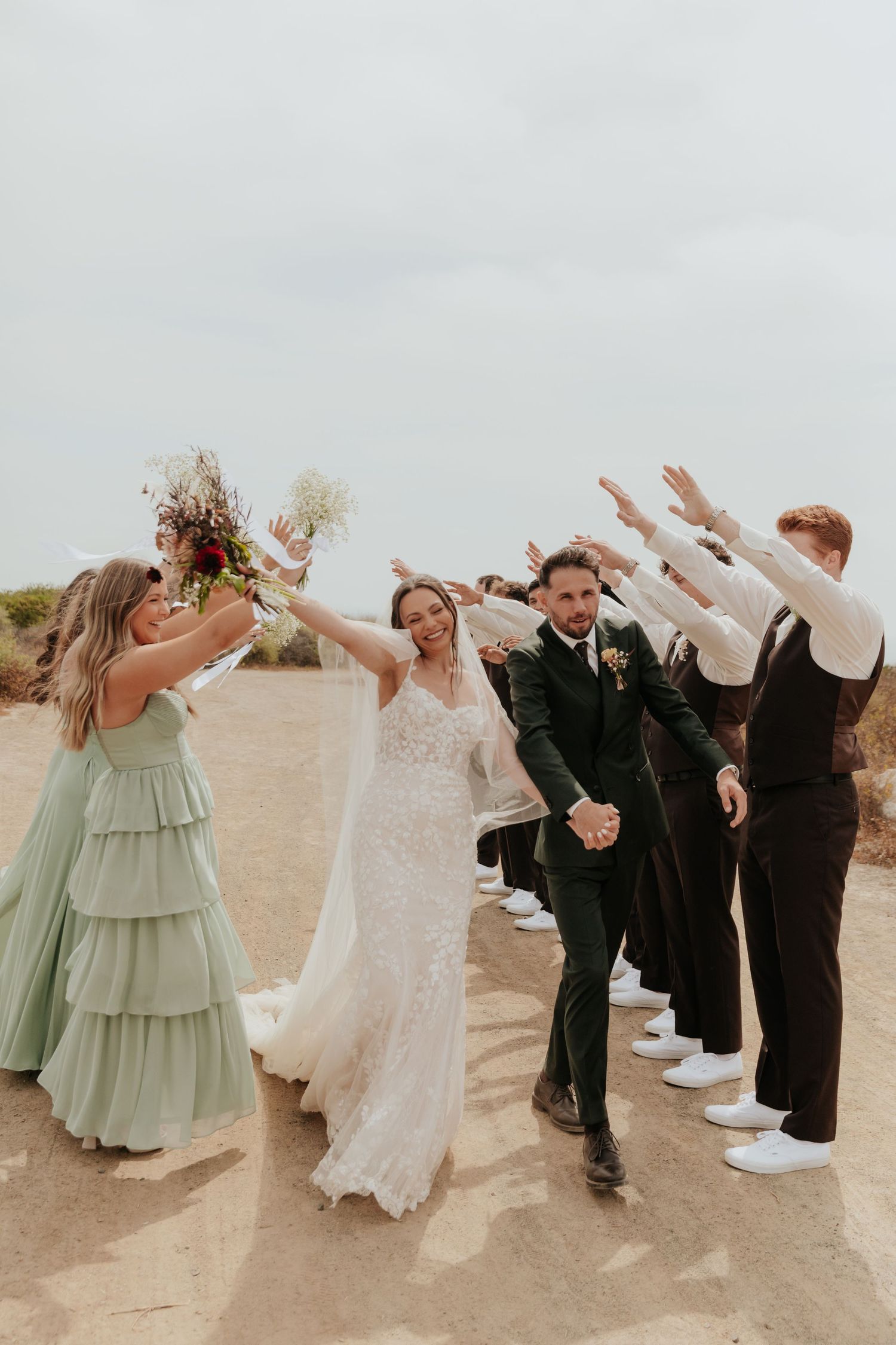 Bride and groom walk through celebratory arch of raised hands formed by wedding party in sage and black attire.