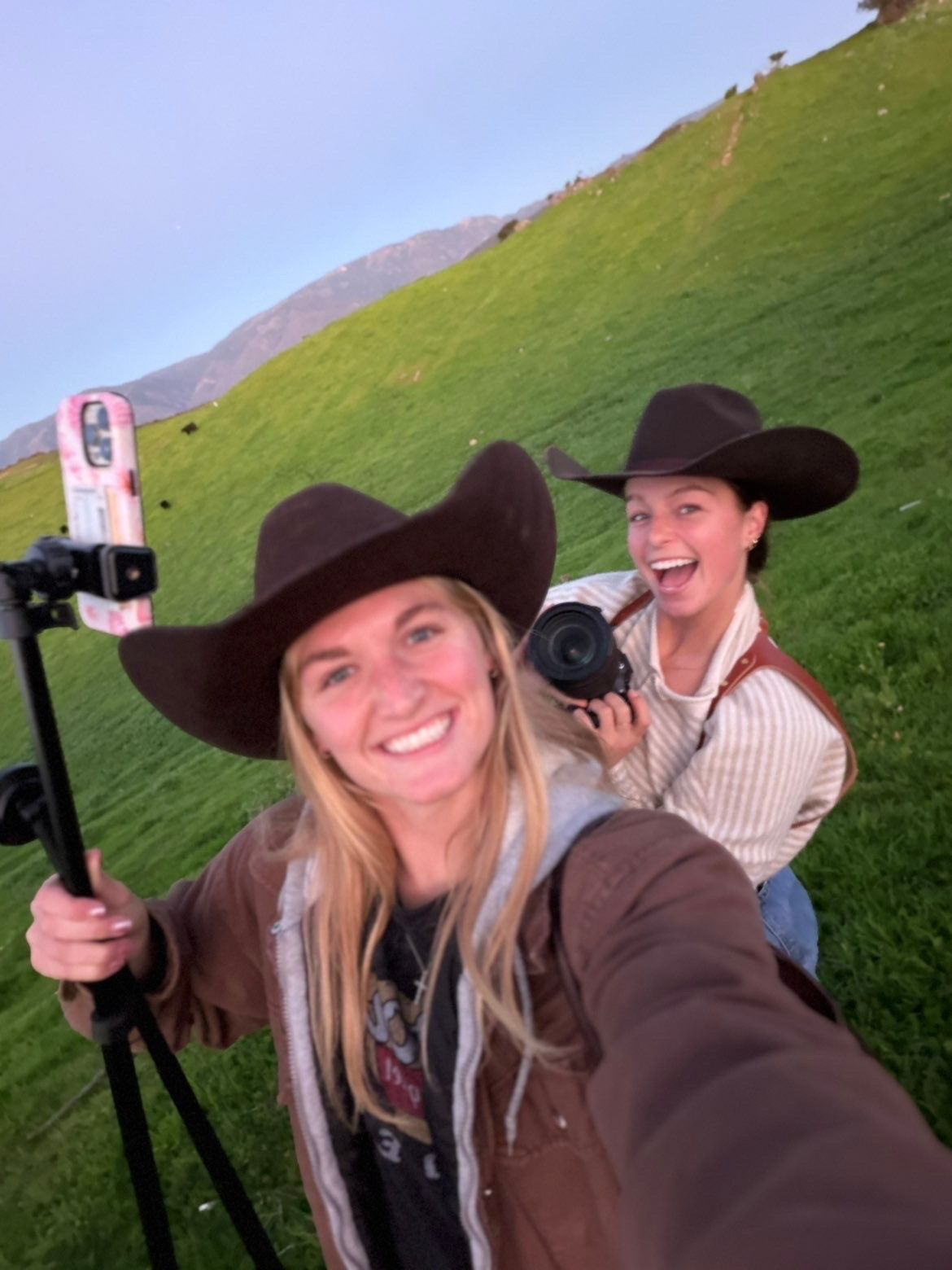 Two women in cowboy hats taking a selfie with cameras in a green mountain meadow.