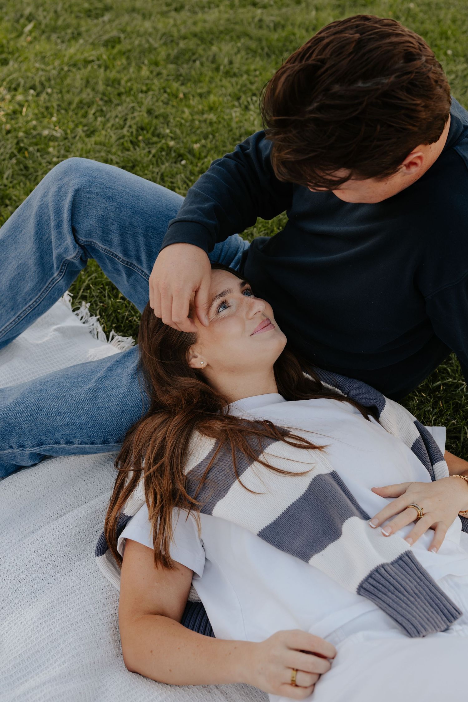 Couple relaxing on blanket in park, woman lying down smiling up at man who gently brushes her hair.
