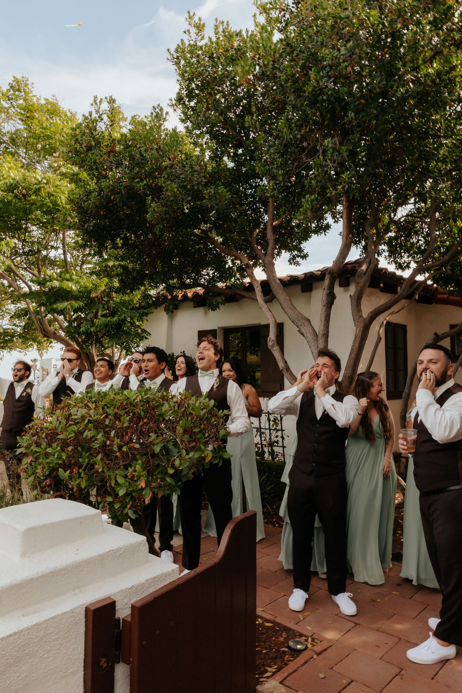 Wedding party in sage green and black attire cheering outdoors near white building surrounded by lush trees.