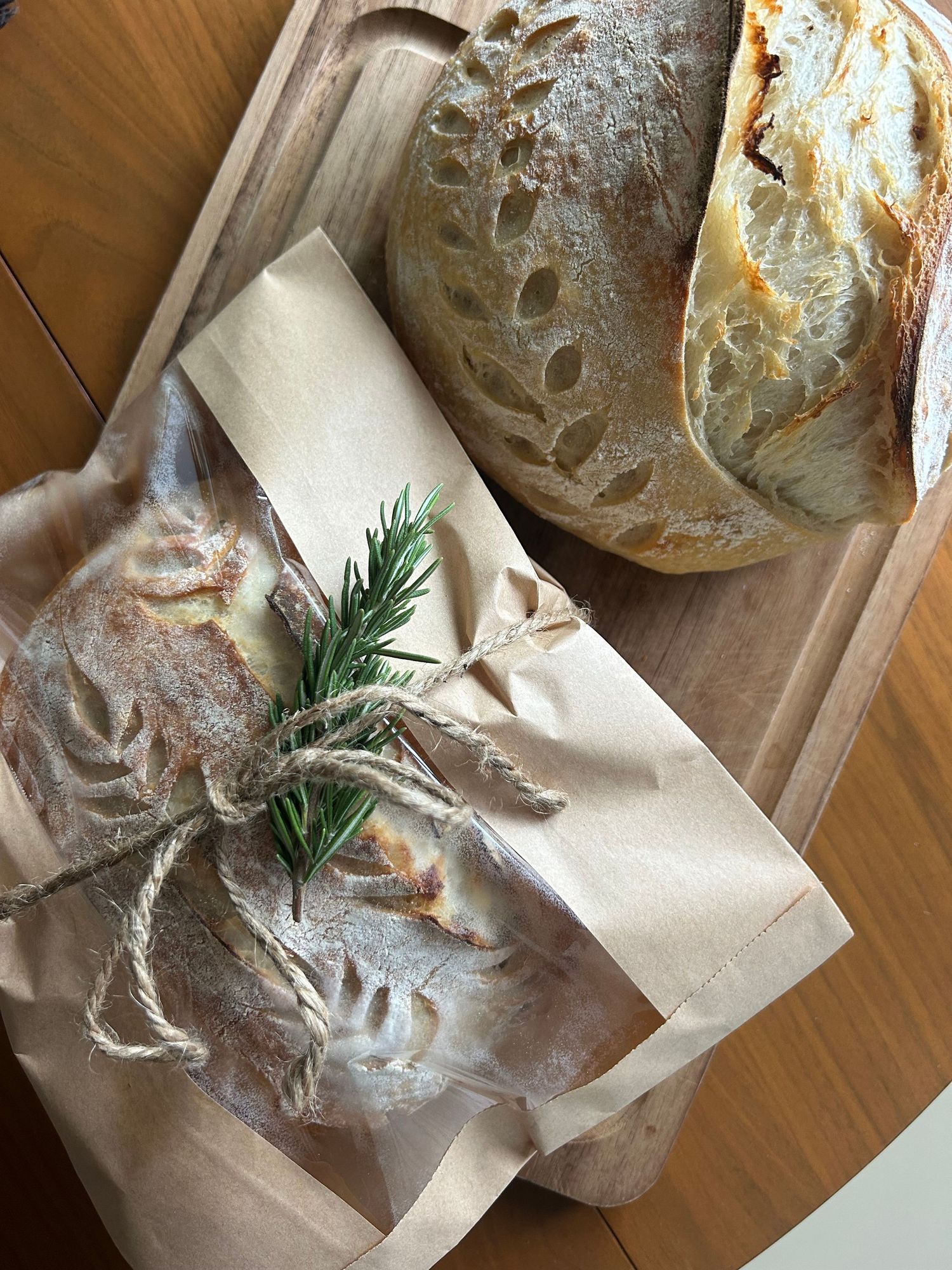 Two rustic sourdough loaves on a wooden board, one wrapped in parchment paper tied with twine and fresh rosemary.