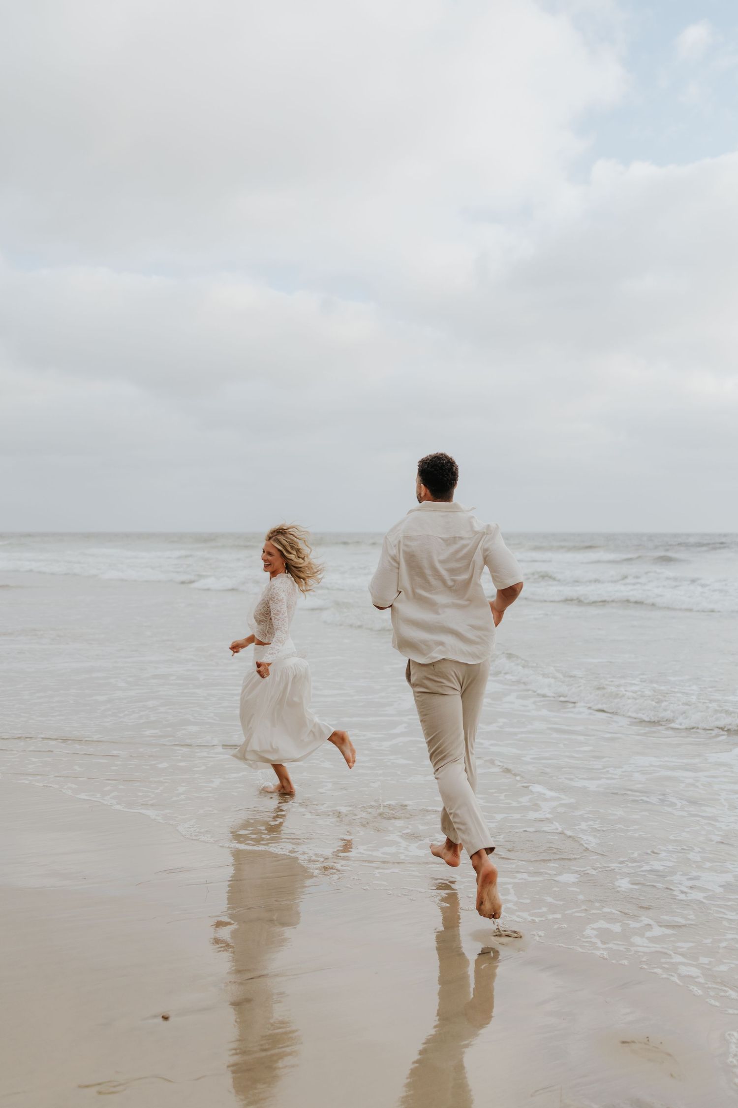 Barefoot couple in white outfits runs playfully along ocean shoreline under cloudy sky.
