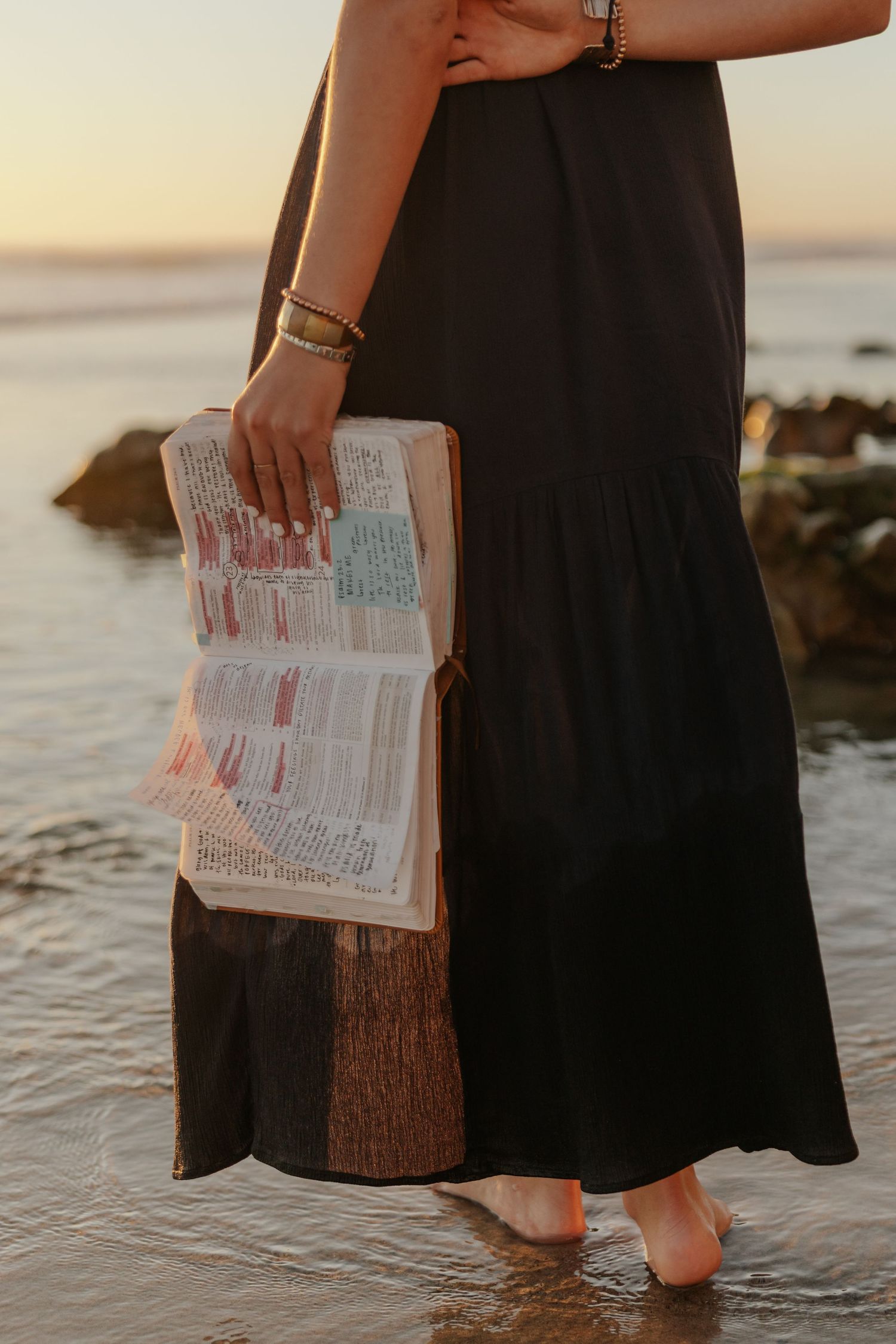 Woman in black maxi dress standing barefoot in ocean water holding an open annotated Bible at sunset.