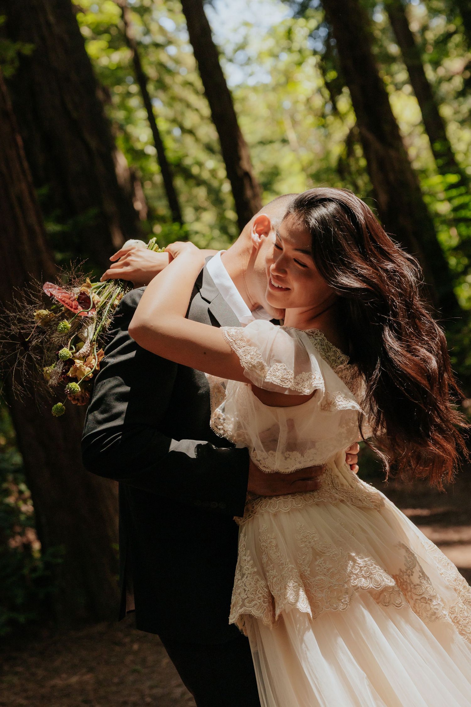 Bride and groom dancing joyfully among tall redwood trees, bride in lace gown holding wildflower bouquet.