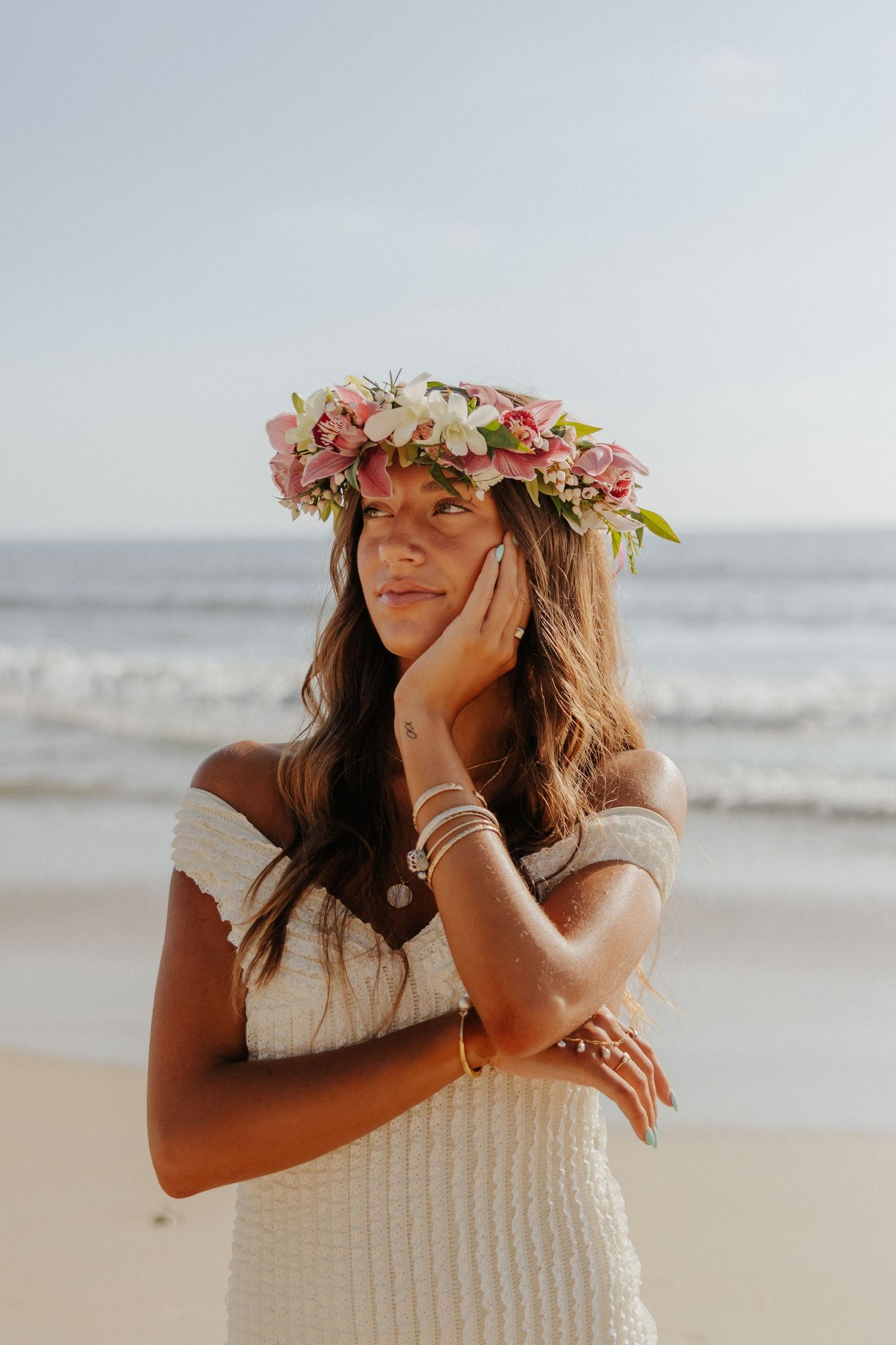 Woman in off-shoulder white dress wears pink and white floral crown on sunny beach shoreline.